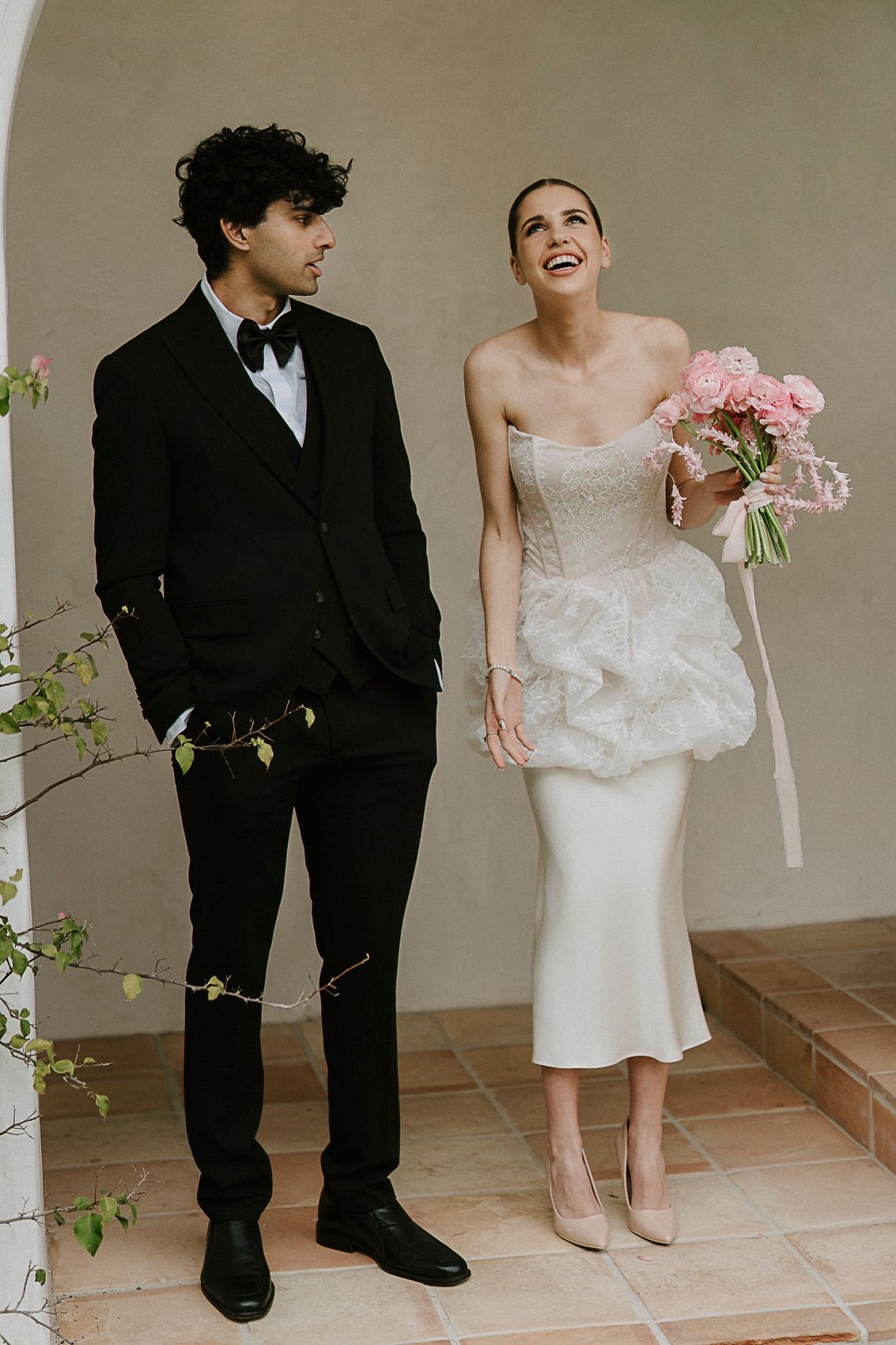 Bride in strapless white midi dress laughs holding pink bouquet beside groom in black tuxedo at outdoor venue.