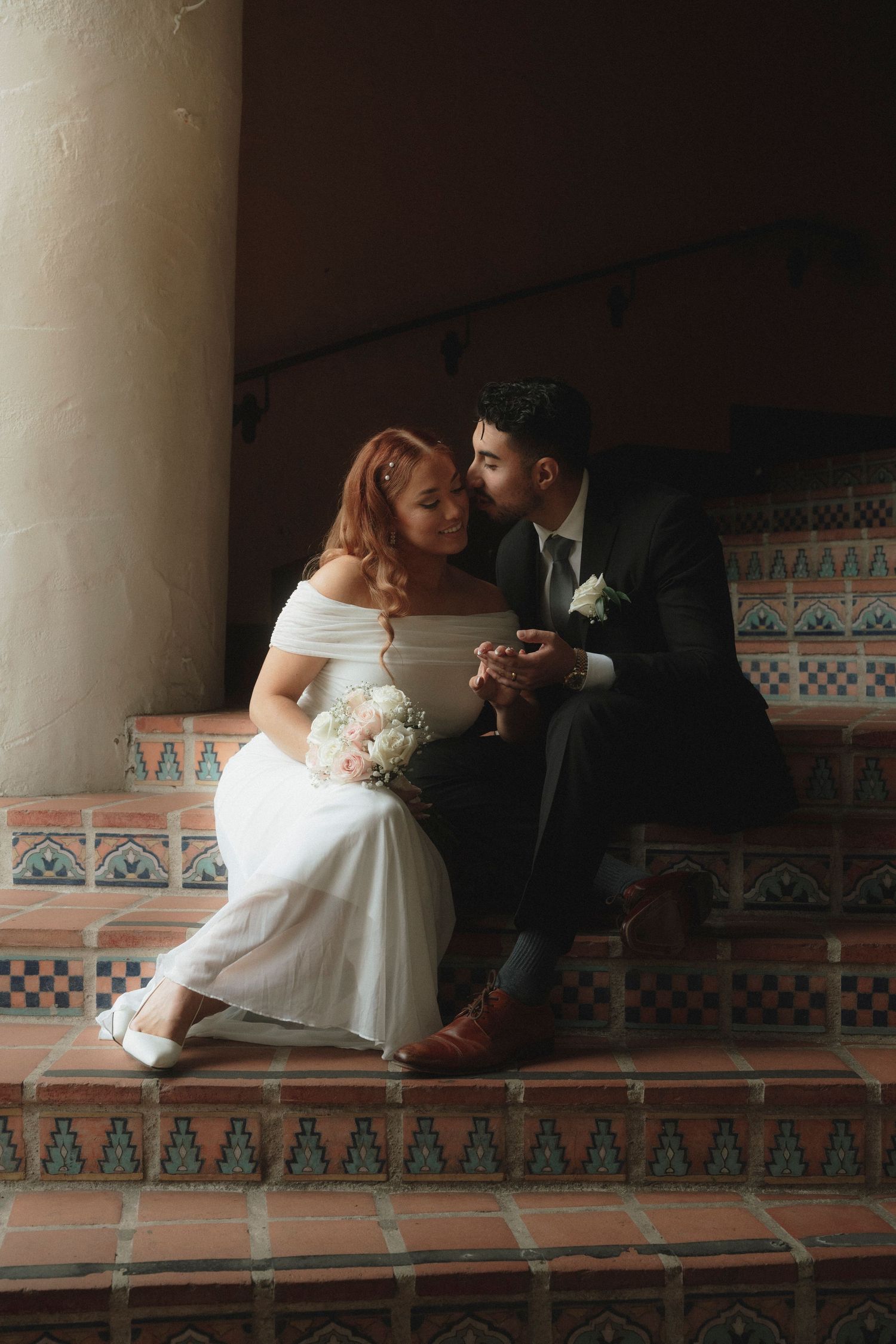 Newlywed couple sitting intimately on decorative tiled steps, bride holding white bouquet in wedding dress, groom in dark suit.