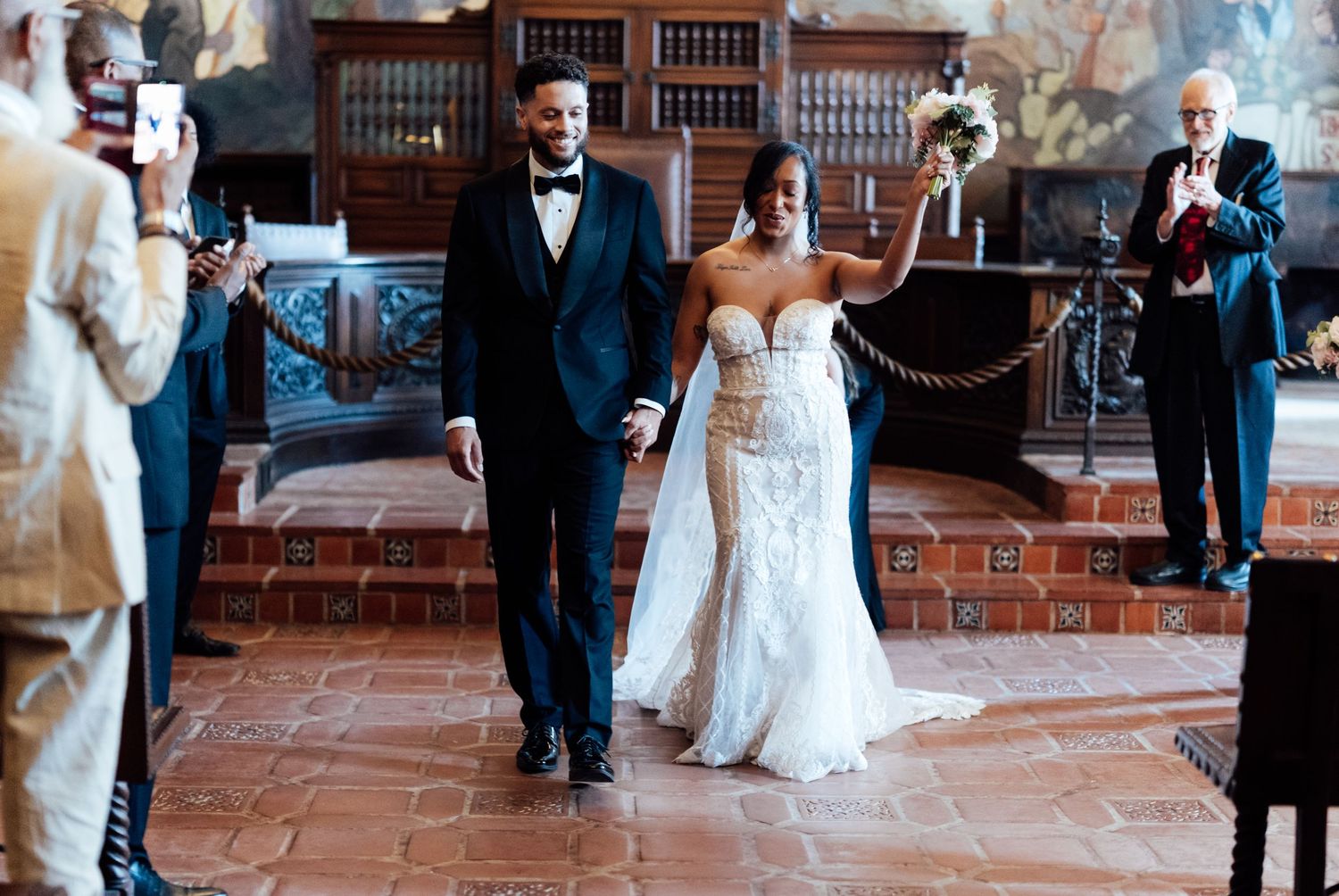 Newlywed couple walks down the aisle in elegant wedding attire inside a grand historic venue.