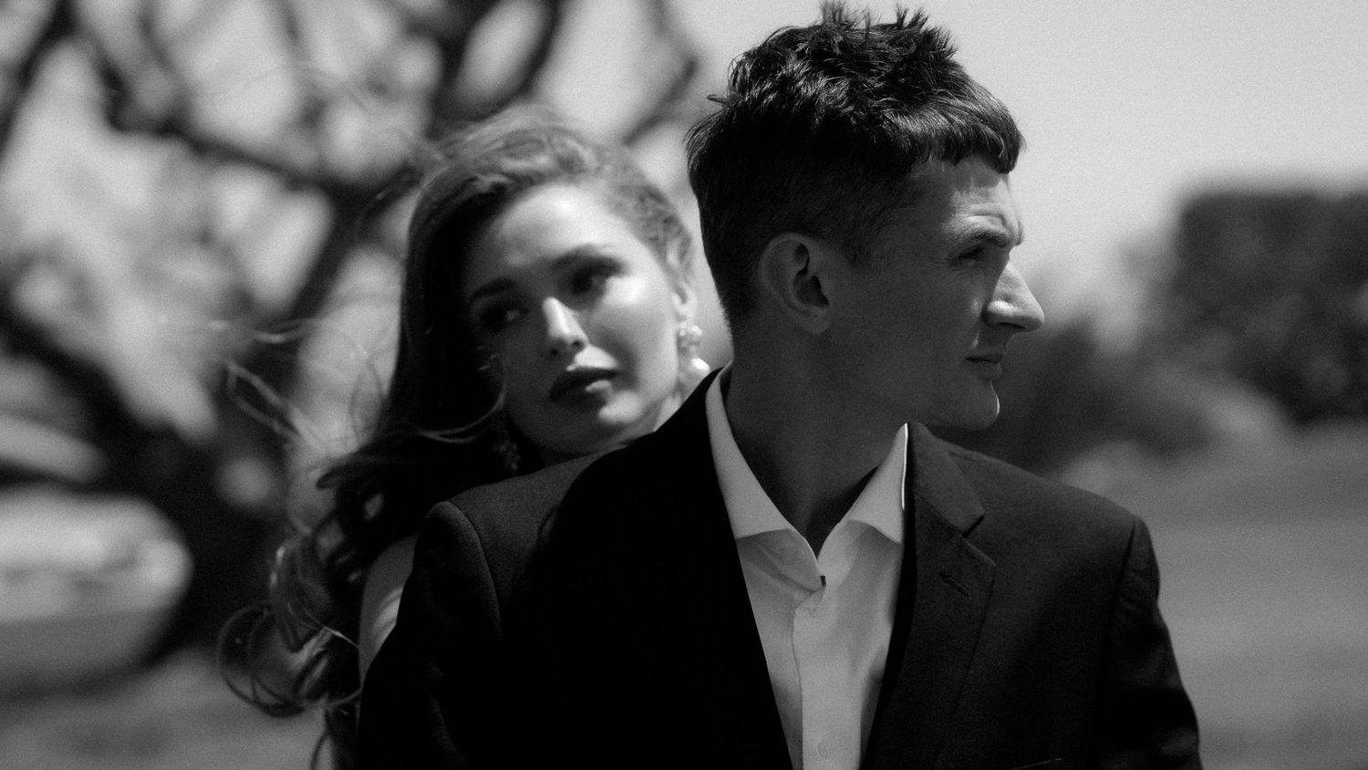 Black and white portrait of a young couple in formal attire standing outdoors near bare trees.
