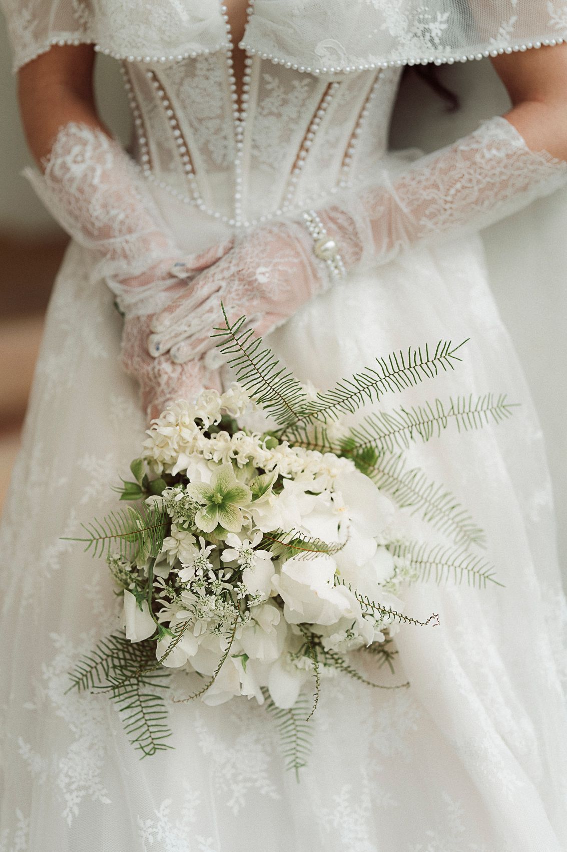 Bride in lace wedding gown and gloves holds elegant white floral bouquet with ferns.