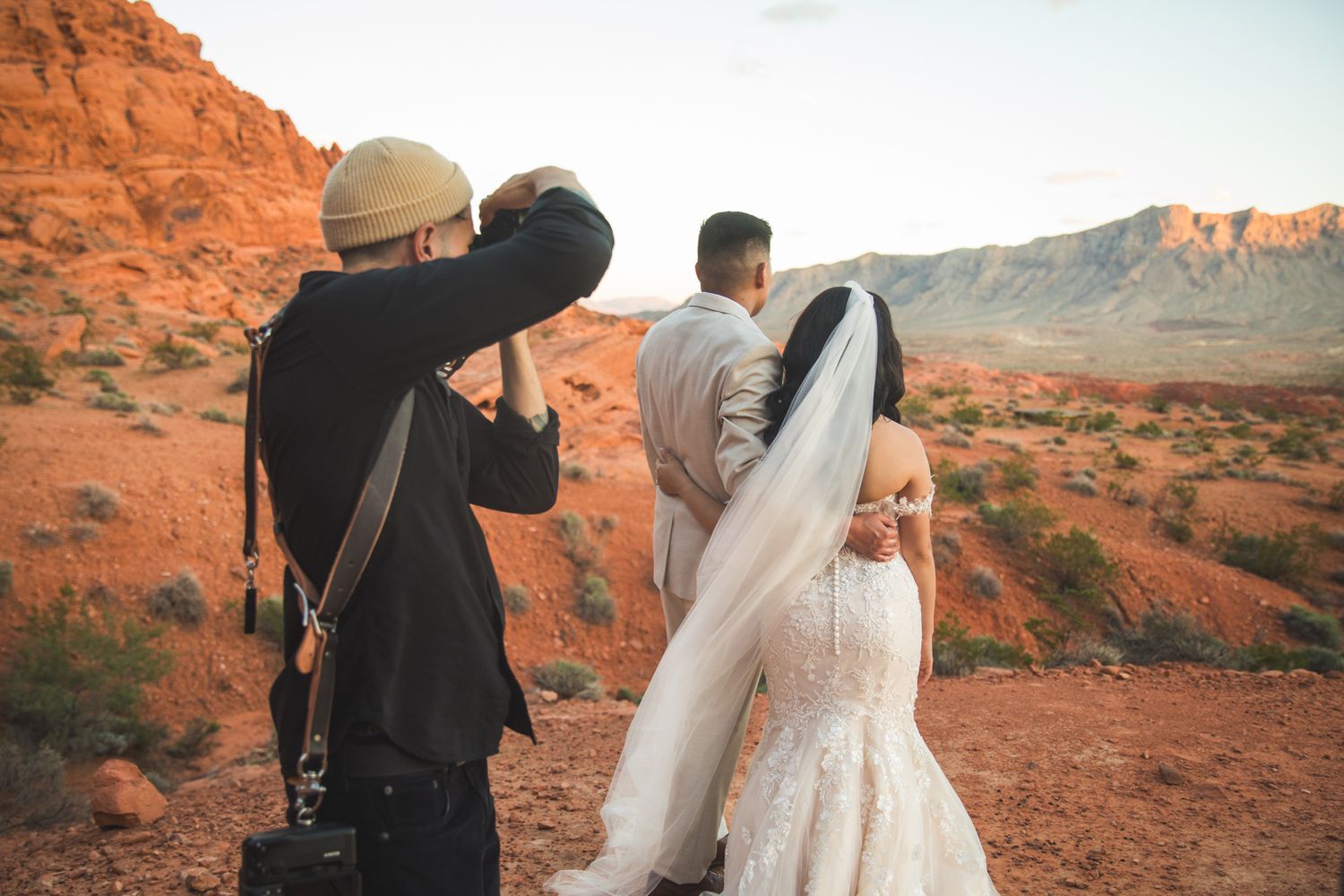 Wedding photographer capturing bride and groom in stunning red rock desert landscape at sunset.