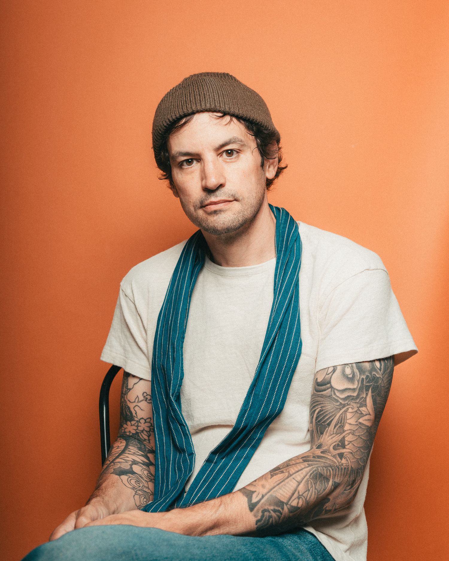 Nick Streeter in brown beanie and blue striped scarf sits casually against orange background.