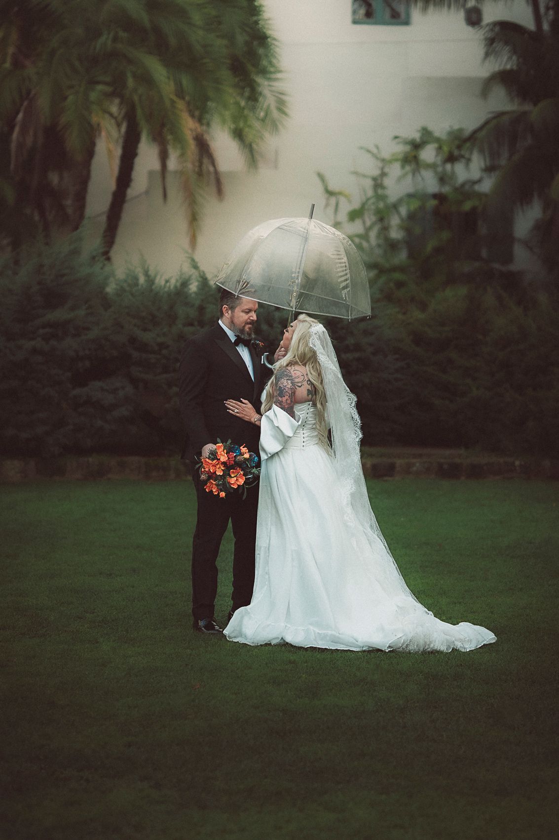 Bride and groom share romantic moment under clear umbrella on lush green lawn at rainy wedding.