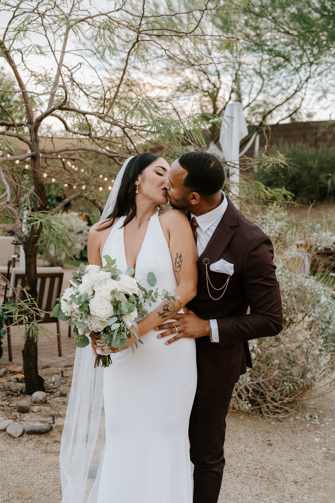 Bride and groom share a kiss outdoors, bride holding white floral bouquet in desert setting.