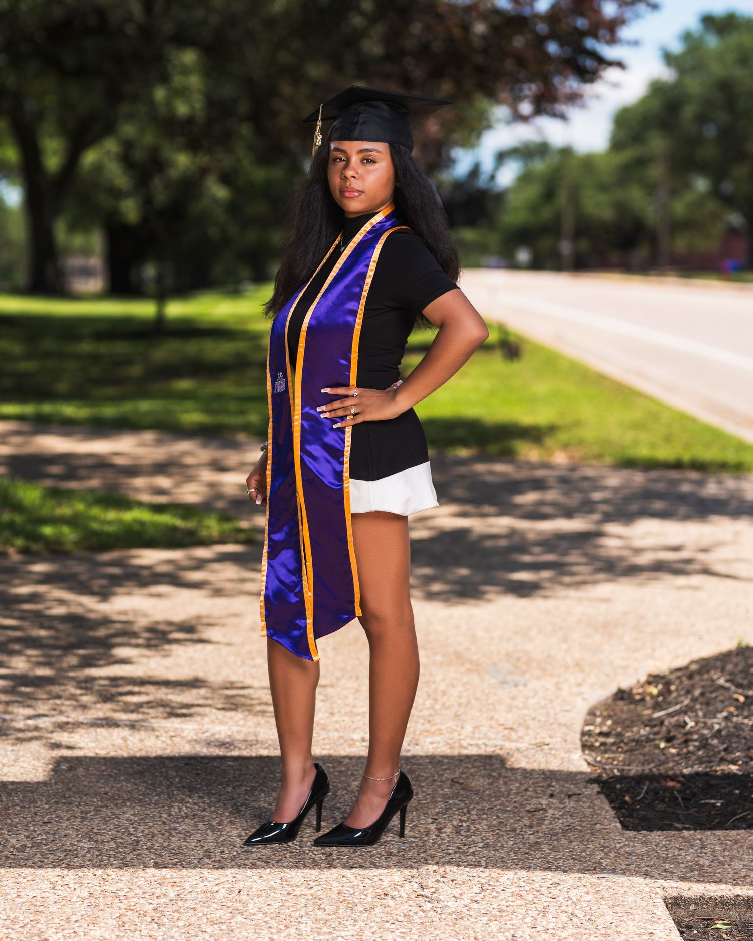 Young woman in graduation cap, purple stole, and heels poses proudly on a tree-lined road for her graduation photo.