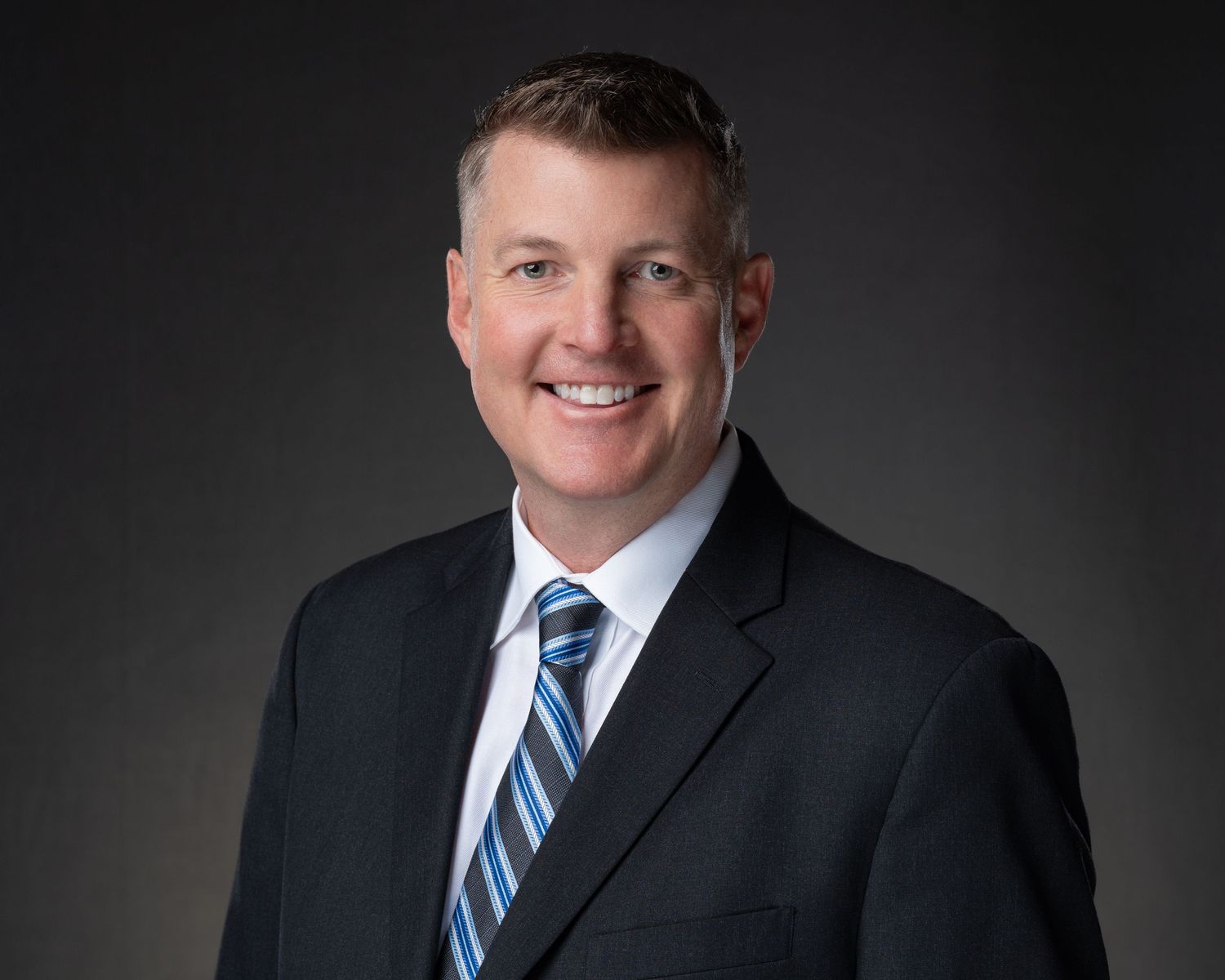 Professional headshot of a smiling man in a dark suit and striped tie against a gray background.