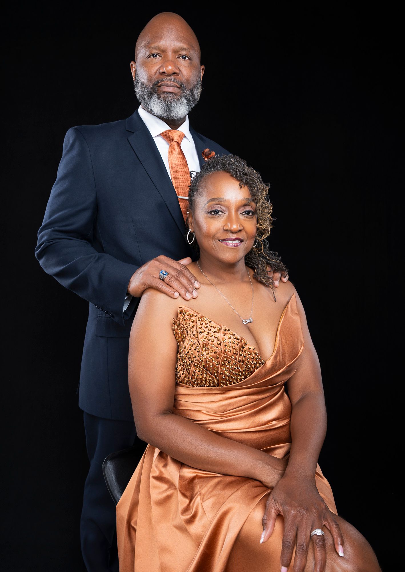 Elegant couple in formal attire posing for portrait against dark background, man standing behind seated woman.