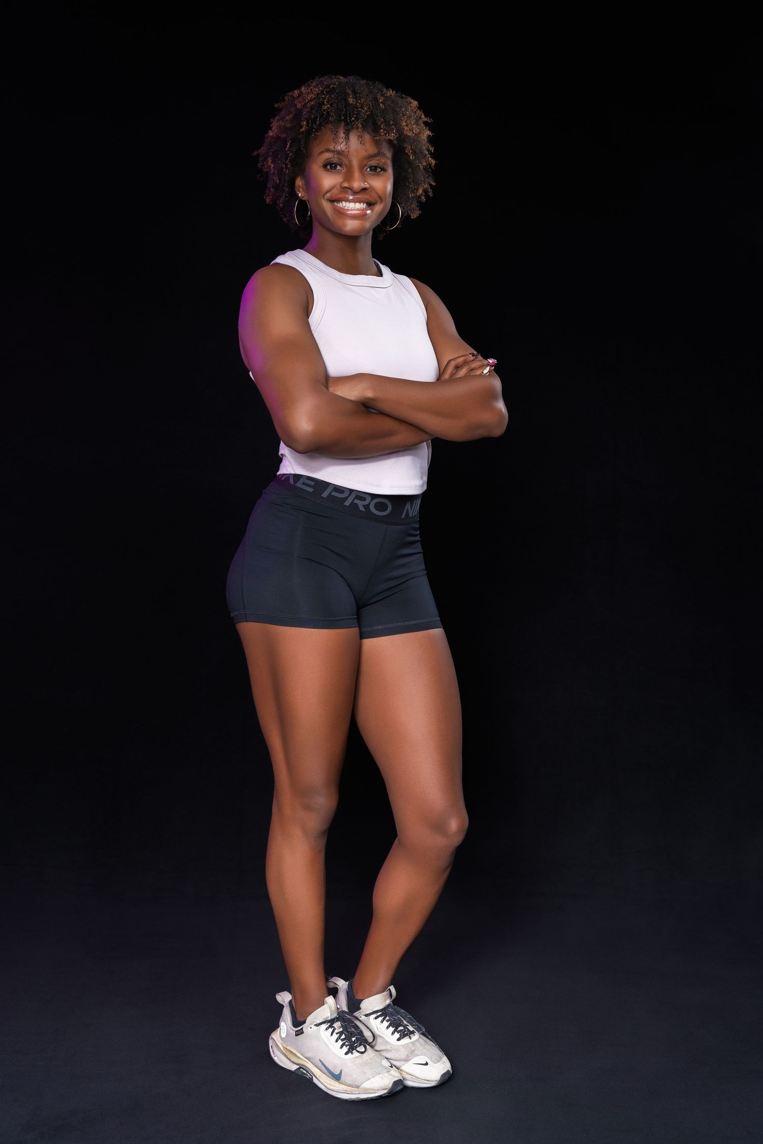 A fit Black woman in white tank and Nike Pro shorts smiles with arms crossed against a black background.