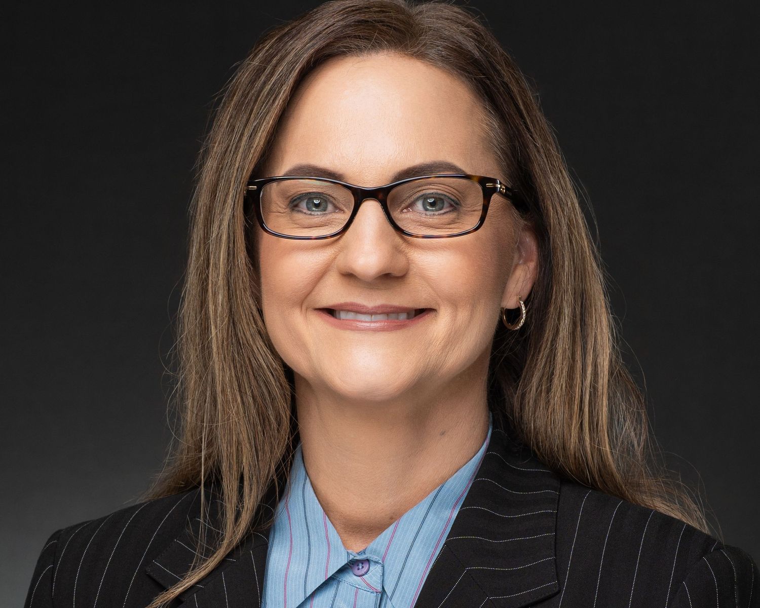 Professional headshot of a woman with glasses and long brown hair wearing a dark blazer and light blue shirt.
