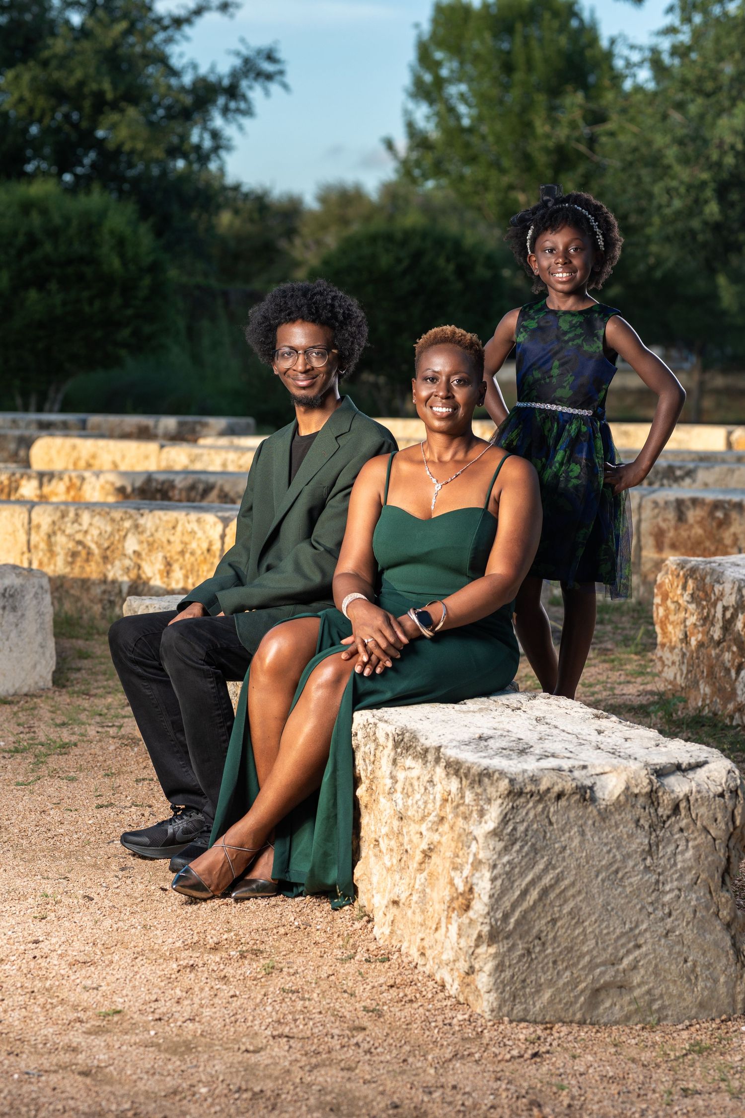 A joyful Black family of three poses on stone ruins outdoors, dressed in elegant green outfits for a formal portrait.