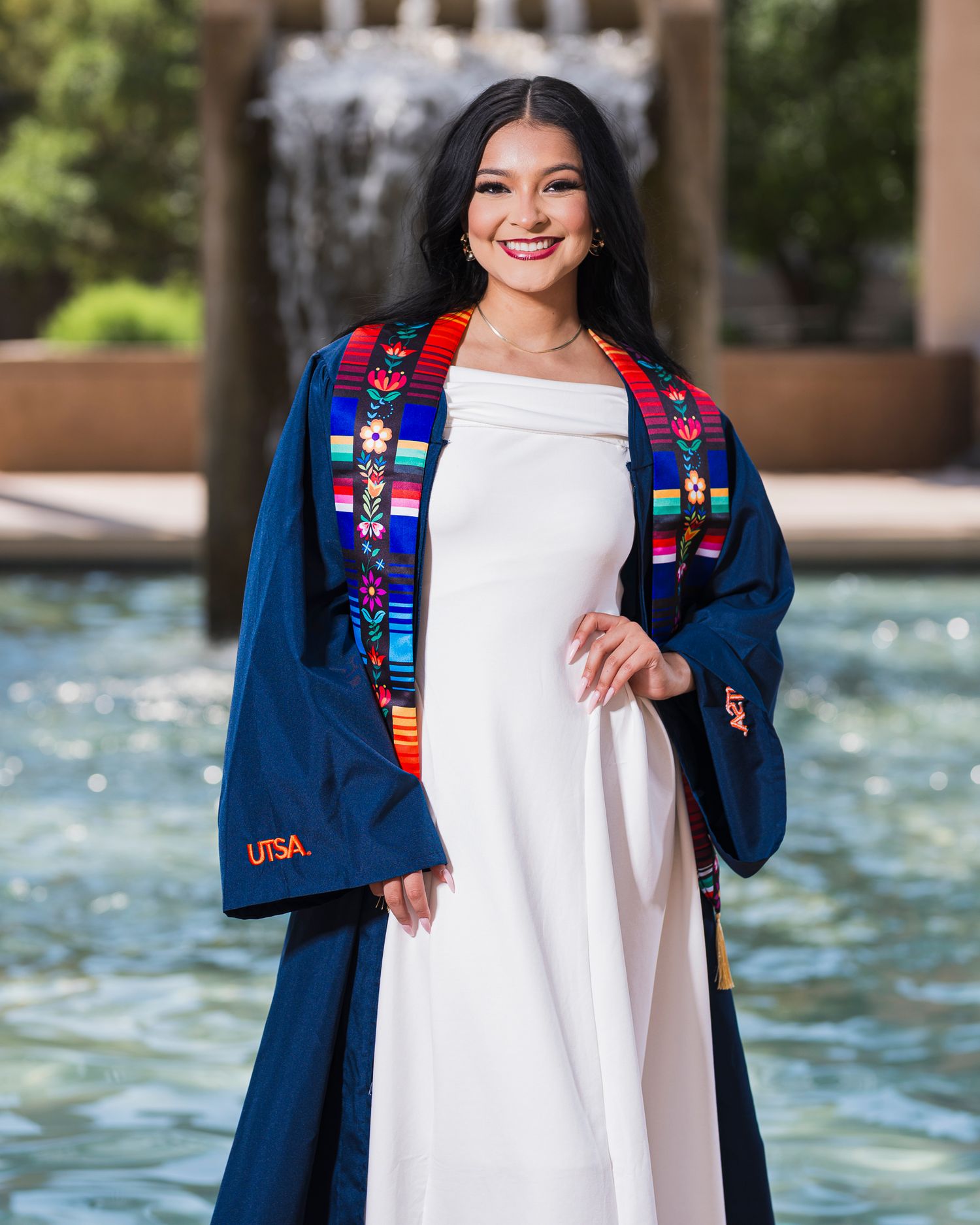 UTSA graduate posing in navy blue graduation gown with multicolored embroidered stole by campus fountain.