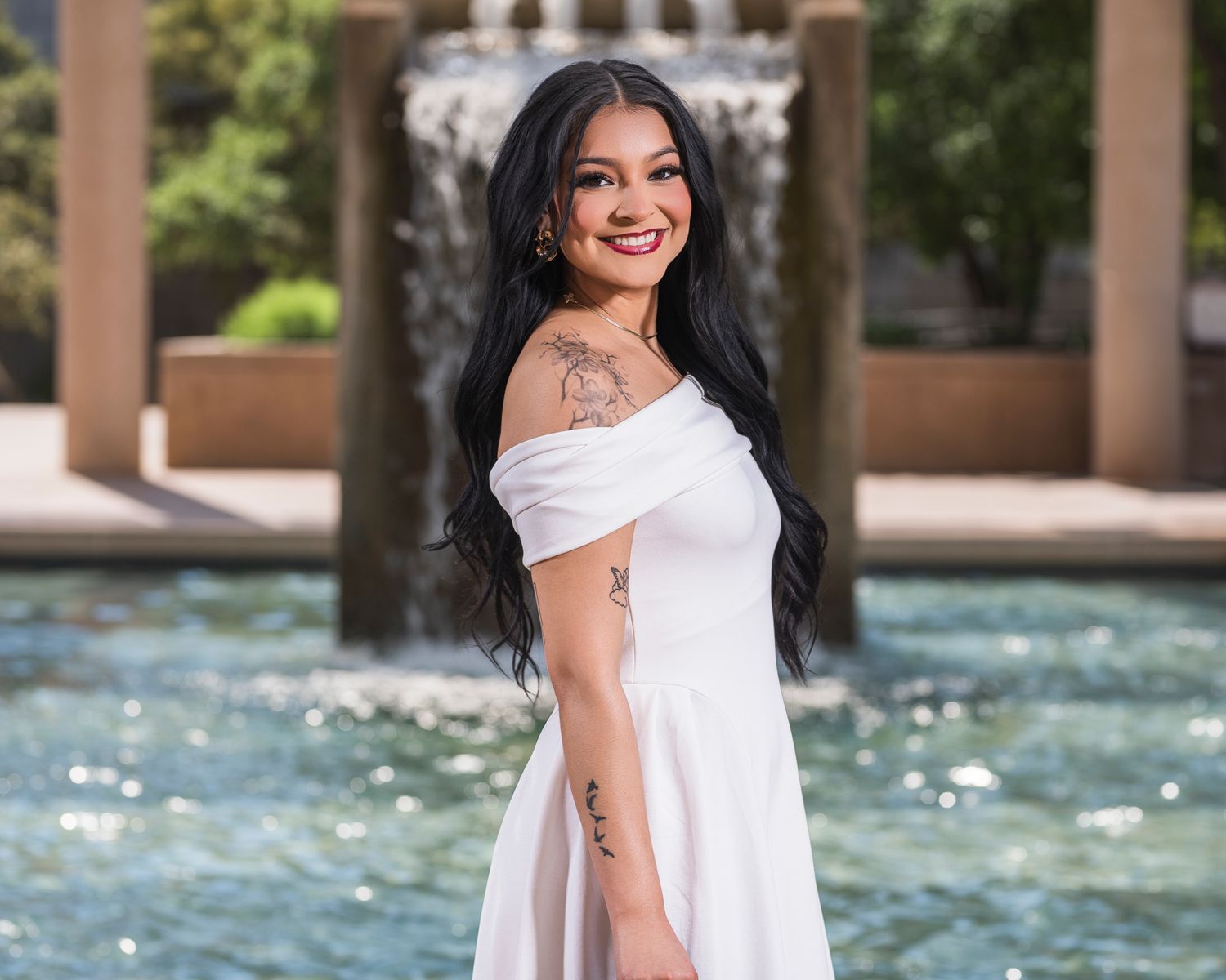 Young woman in white off-shoulder dress smiling by decorative fountain outdoors during a graduation shoot.