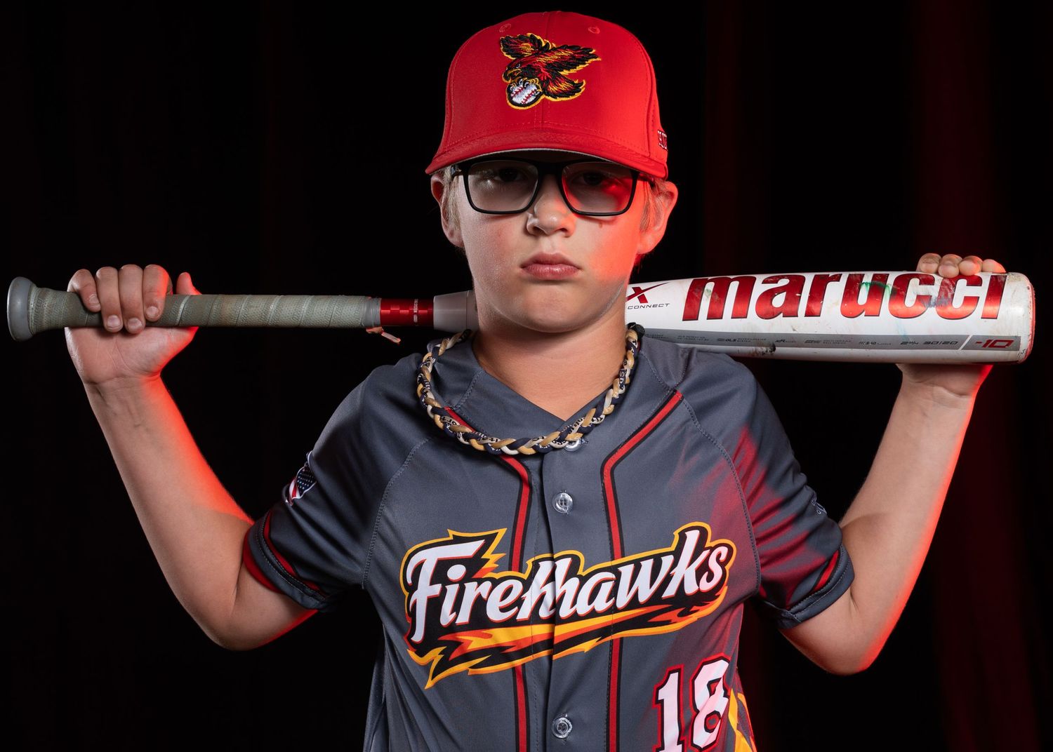 Youth Firehawks baseball player number 18 posing with Marucci bat over shoulder in gray uniform.