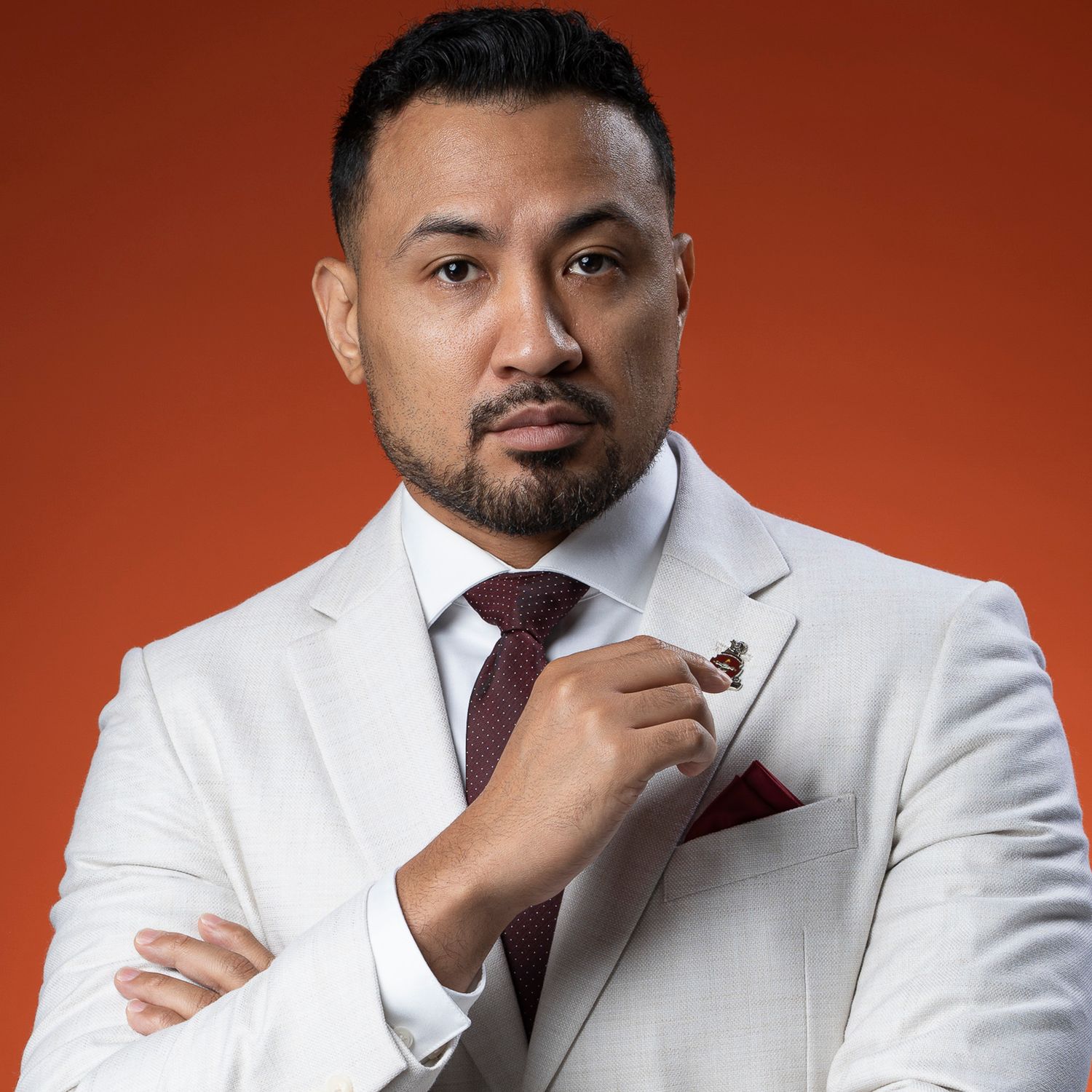 Man in white suit and burgundy tie posing against orange background.