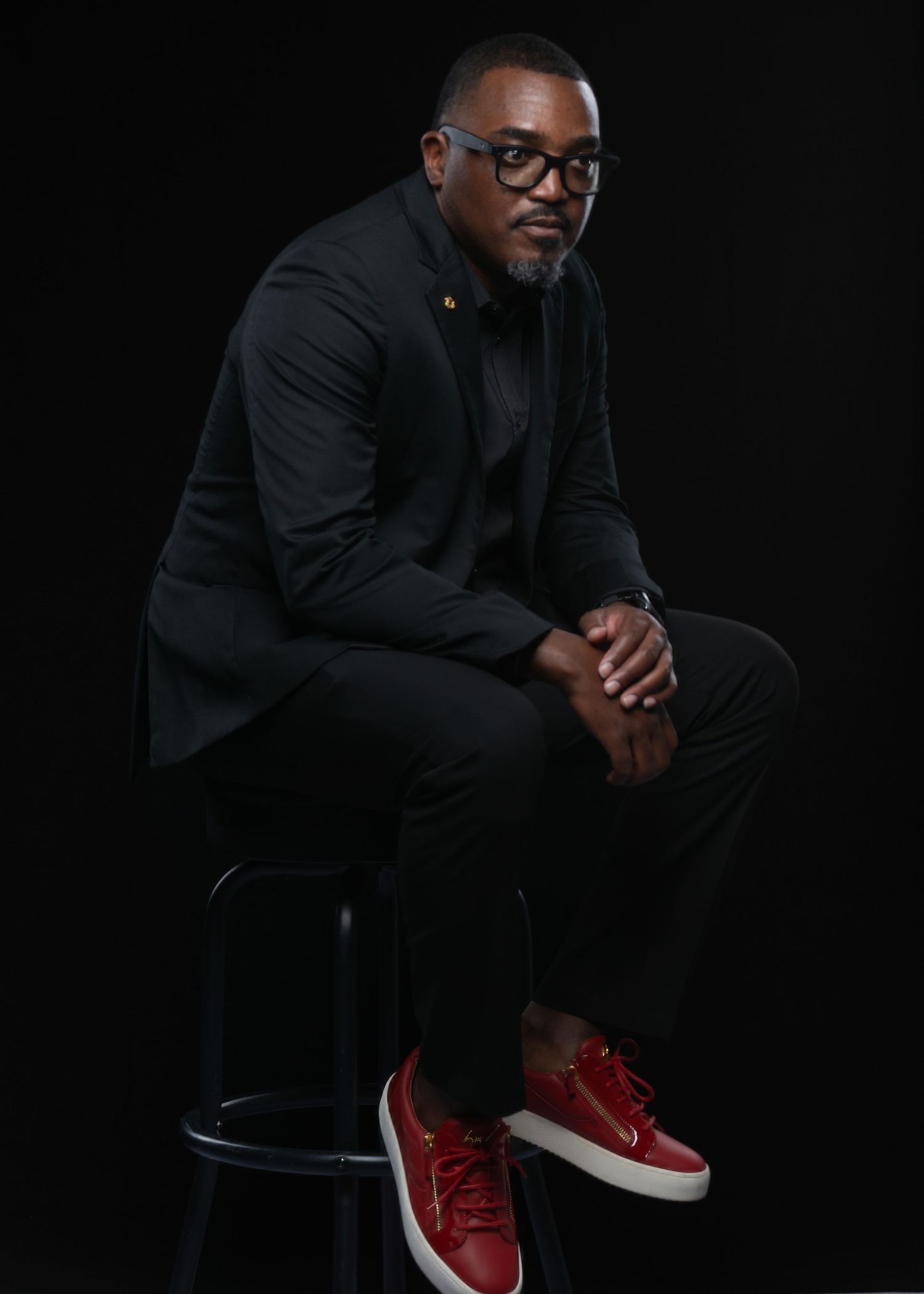 Professional man in black suit and red sneakers sitting on stool against dark background.