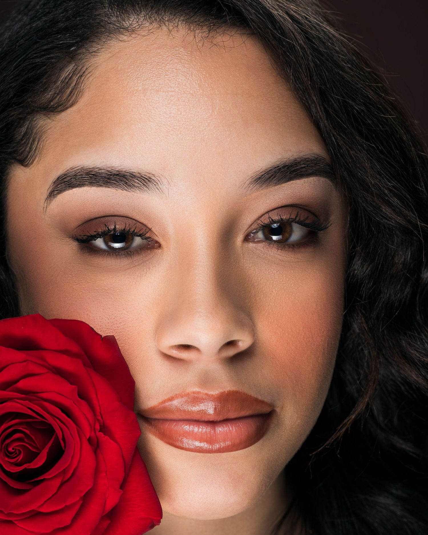 Close-up portrait of young woman with dark eyes holding a vibrant red rose beside her face.