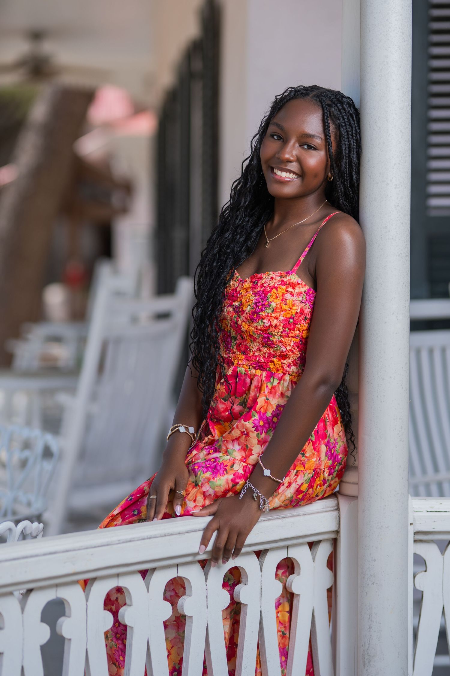 Young woman in vibrant pink floral sundress leaning on white railing with long braided hair.