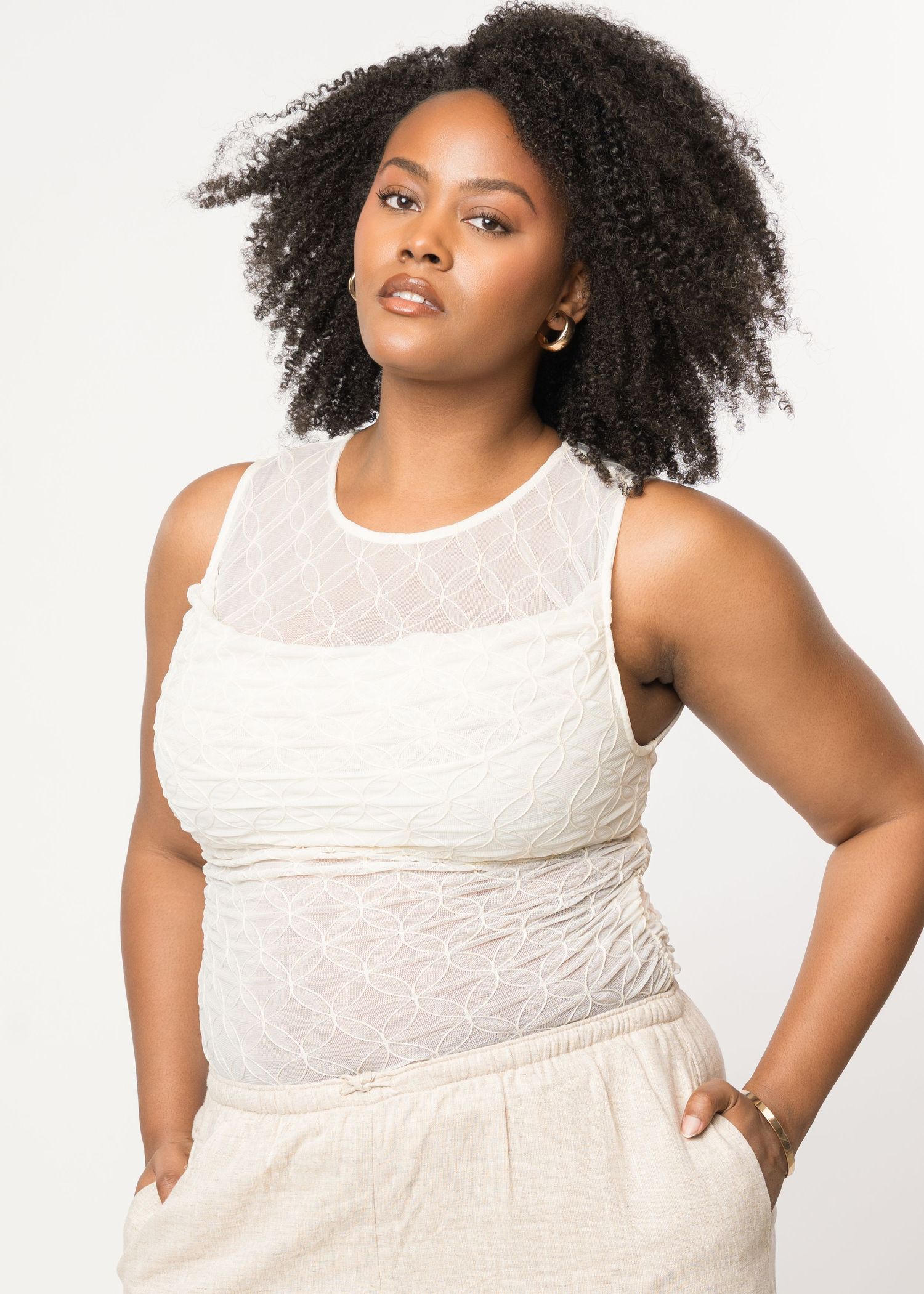 Woman in cream textured sleeveless top posing against white background.