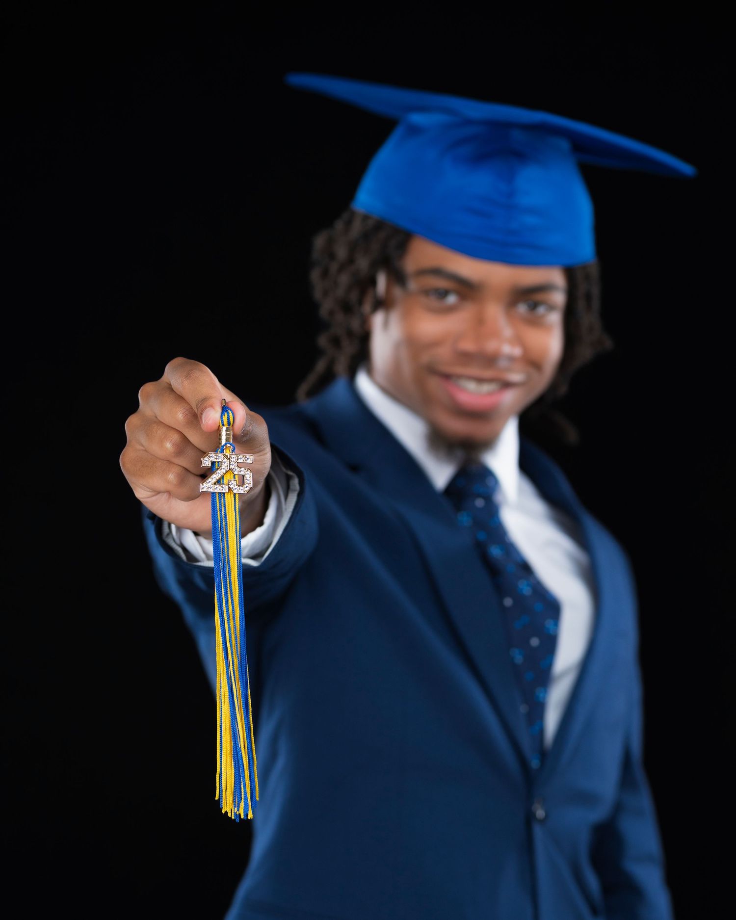 Young man in blue graduation cap and suit proudly holds up a medallion with colorful tassels against black background.