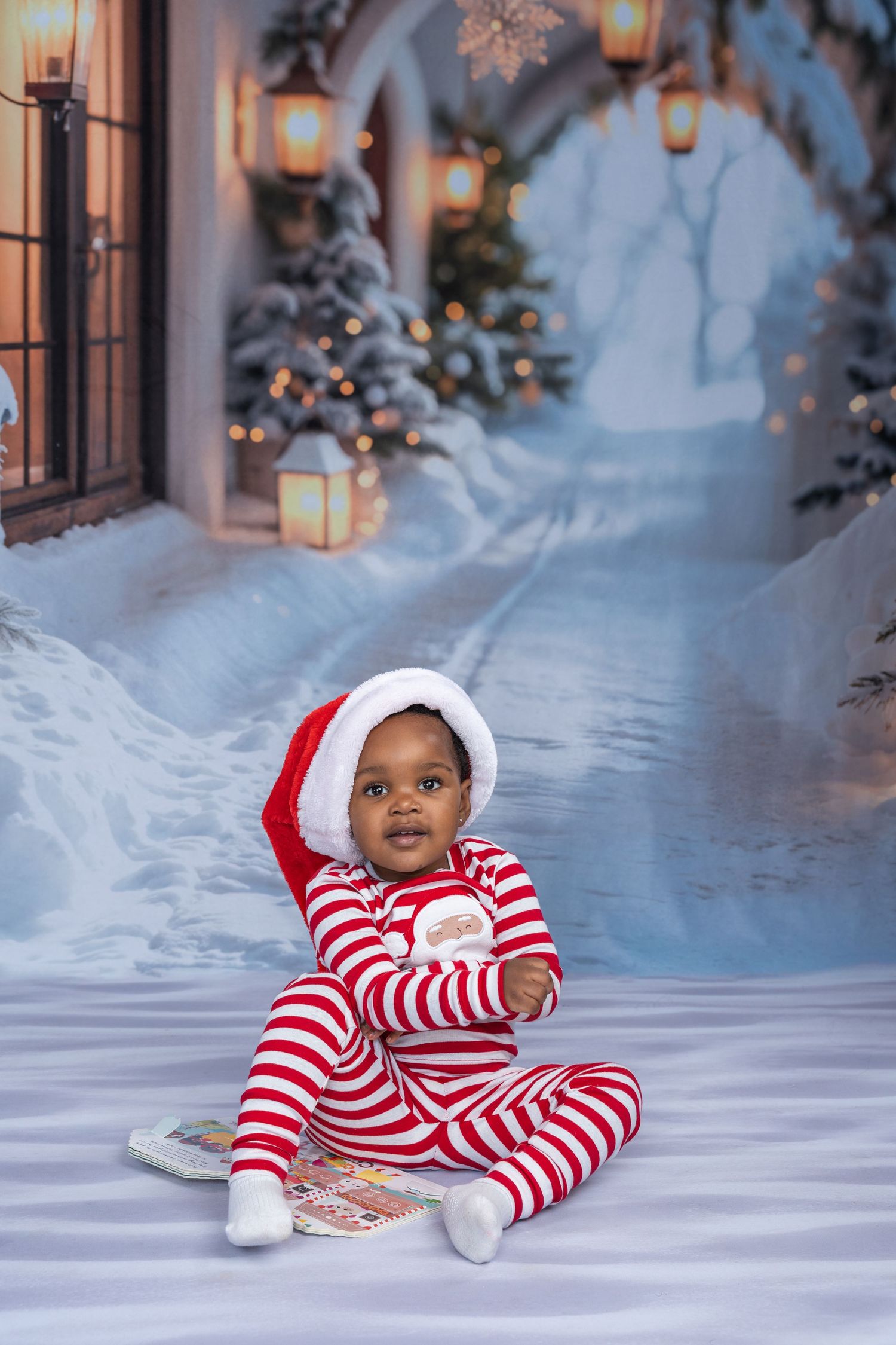 Adorable baby in red striped Santa pajamas and hat sits cross-legged in a festive snowy Christmas backdrop.