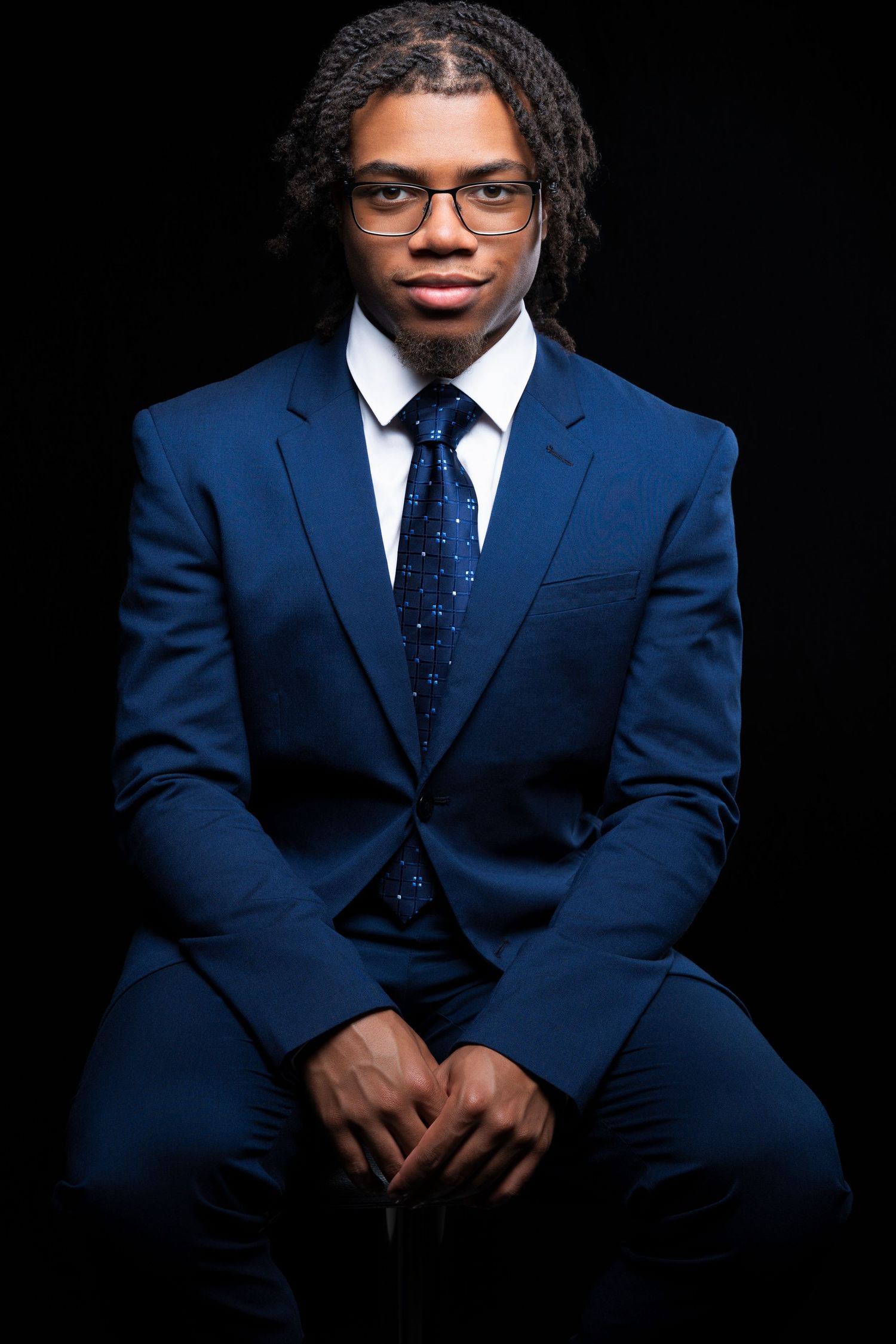 Young man in blue suit, white shirt, tie, and glasses, sitting confidently against black background.
