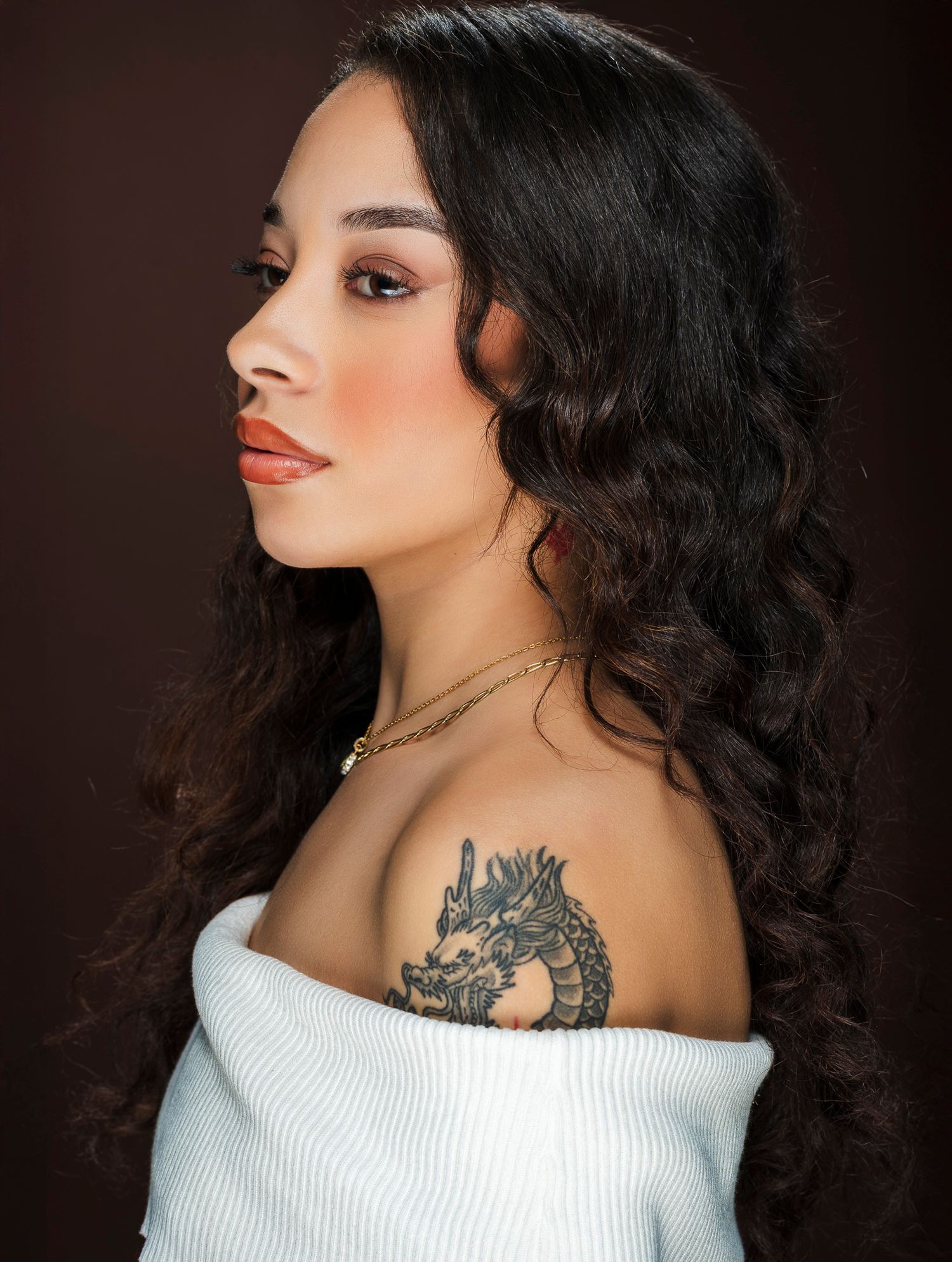 Side profile of woman with dragon shoulder tattoo and long curly hair wearing off-shoulder white top.