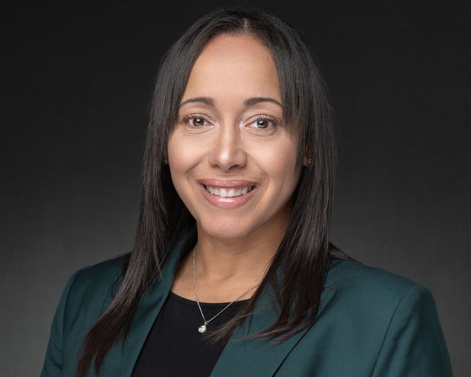 Professional headshot of a woman in a teal blazer smiling against a dark gray background.