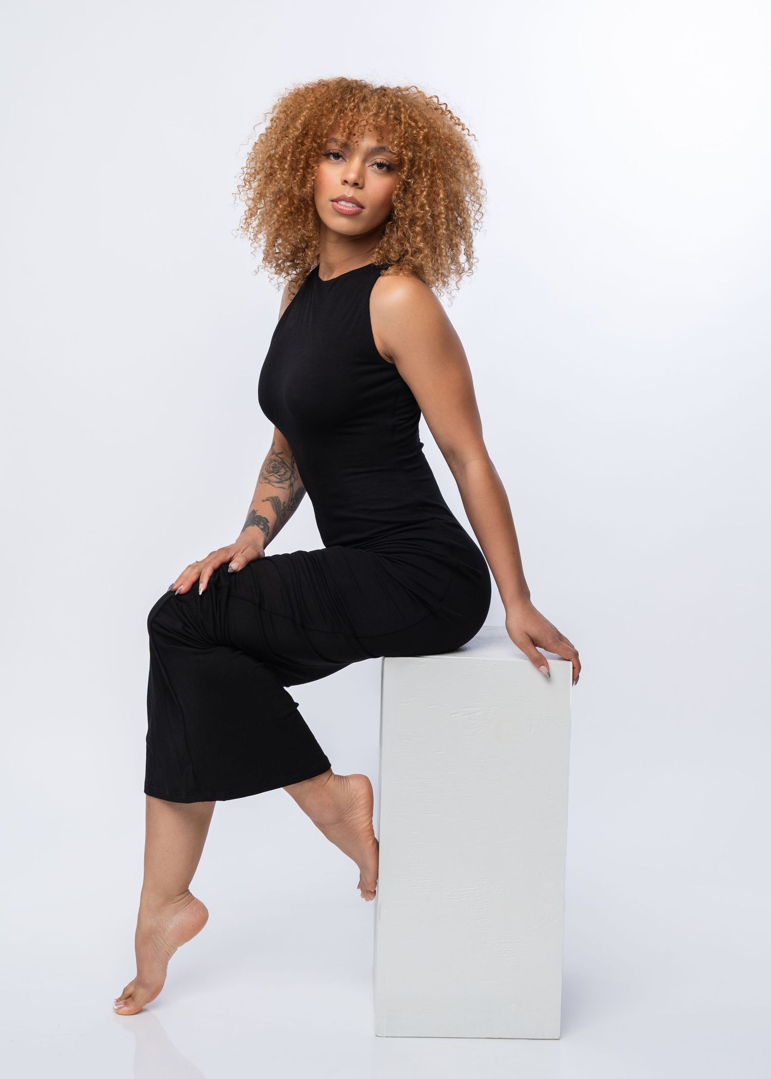 Woman in black sleeveless jumpsuit sitting on white cube, curly auburn hair, tattoo visible, barefoot, white background.