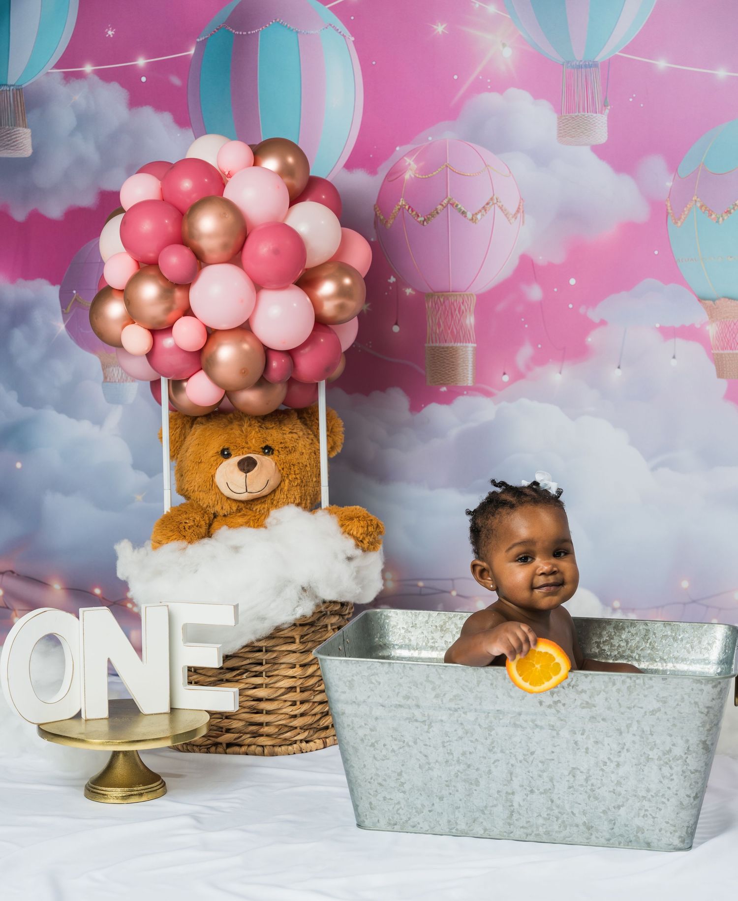 Baby's first birthday cake smash in a tin tub with teddy bear hot air balloon decor and pink cloud backdrop.