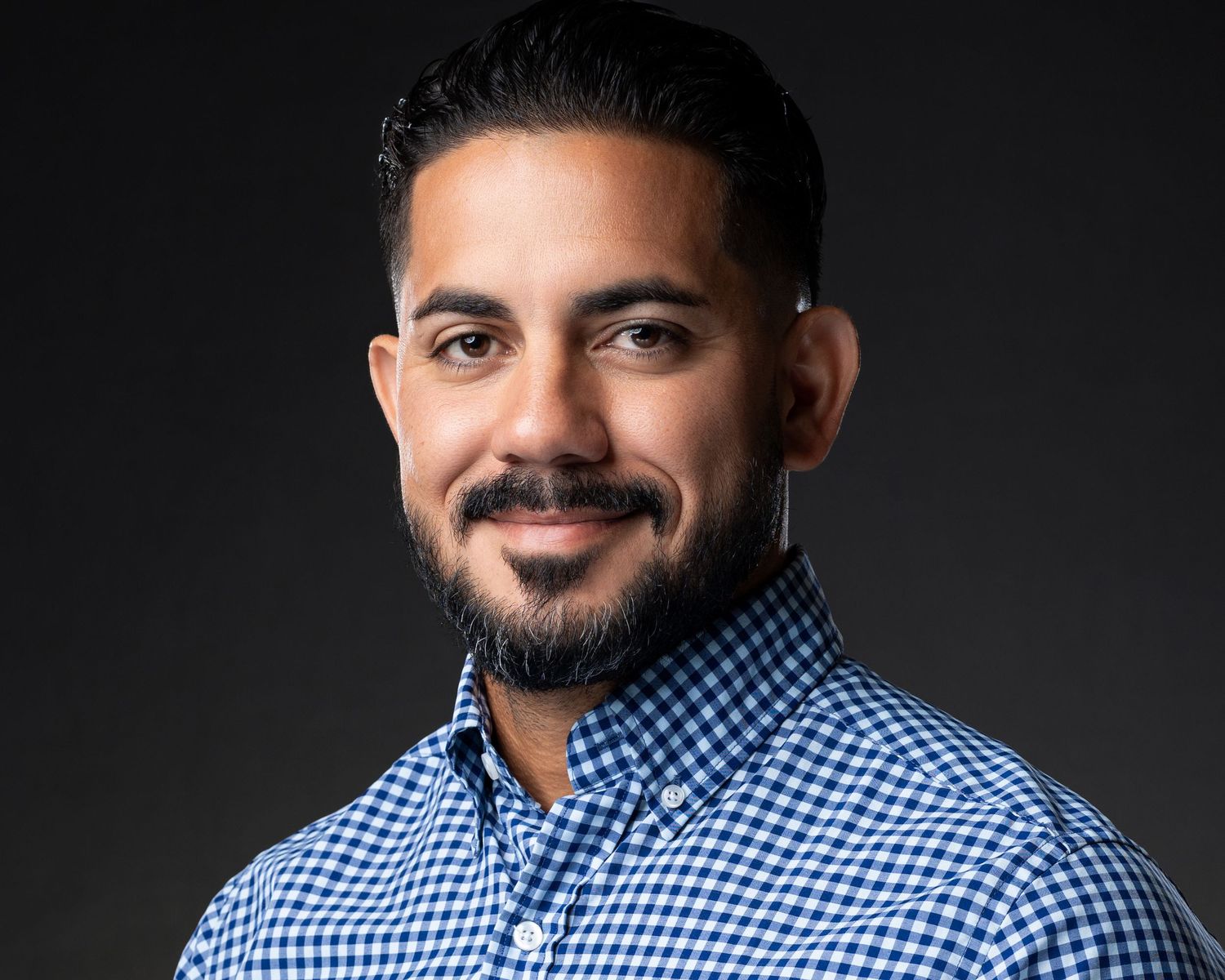 Professional headshot of a smiling man with a beard wearing a blue gingham shirt against a dark background.