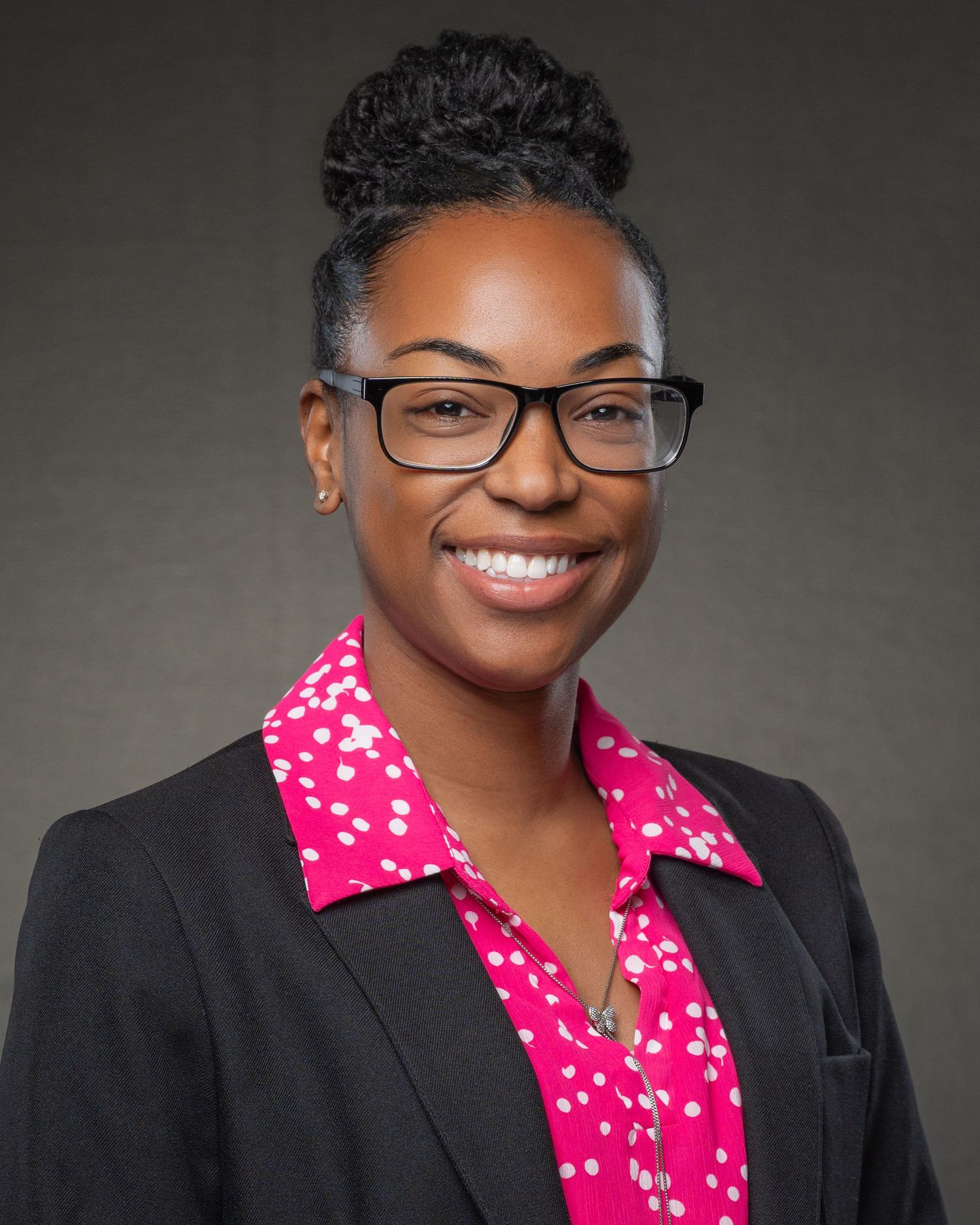 Professional woman in pink polka dot blouse and black blazer wearing glasses, smiling for headshot.