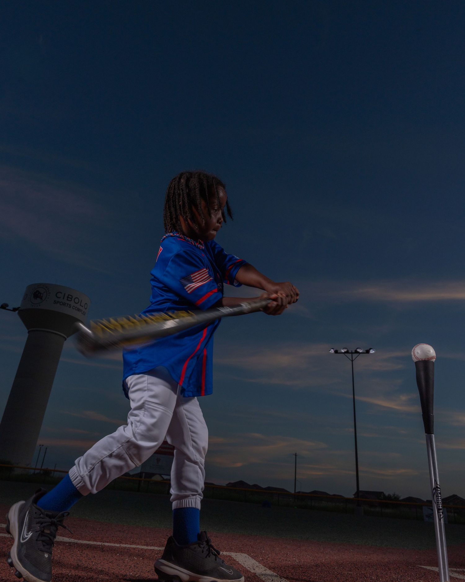 Young baseball player in blue jersey swinging bat at dusk on outdoor baseball field.