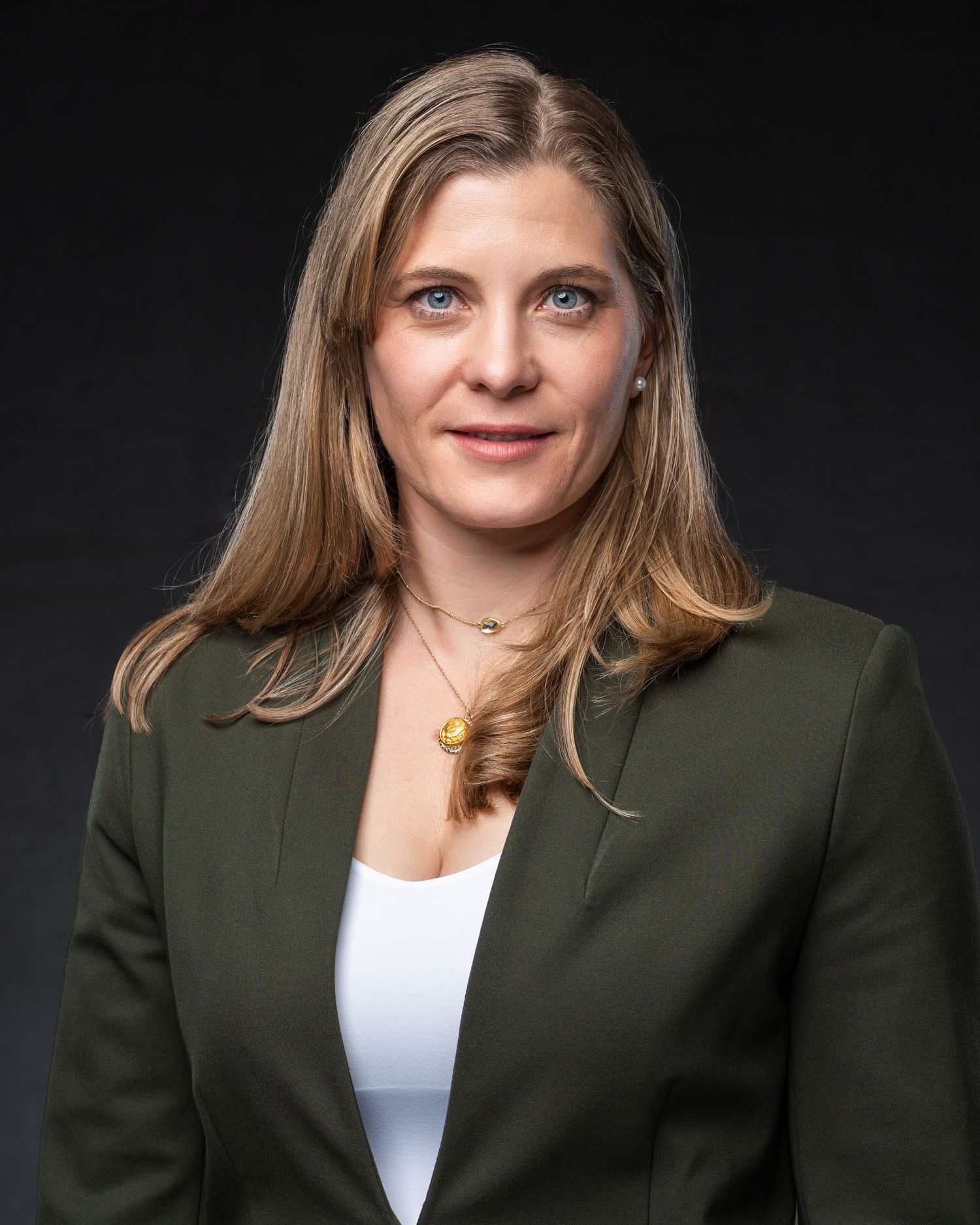Professional woman in green blazer with long blonde hair poses for a corporate headshot against dark background.