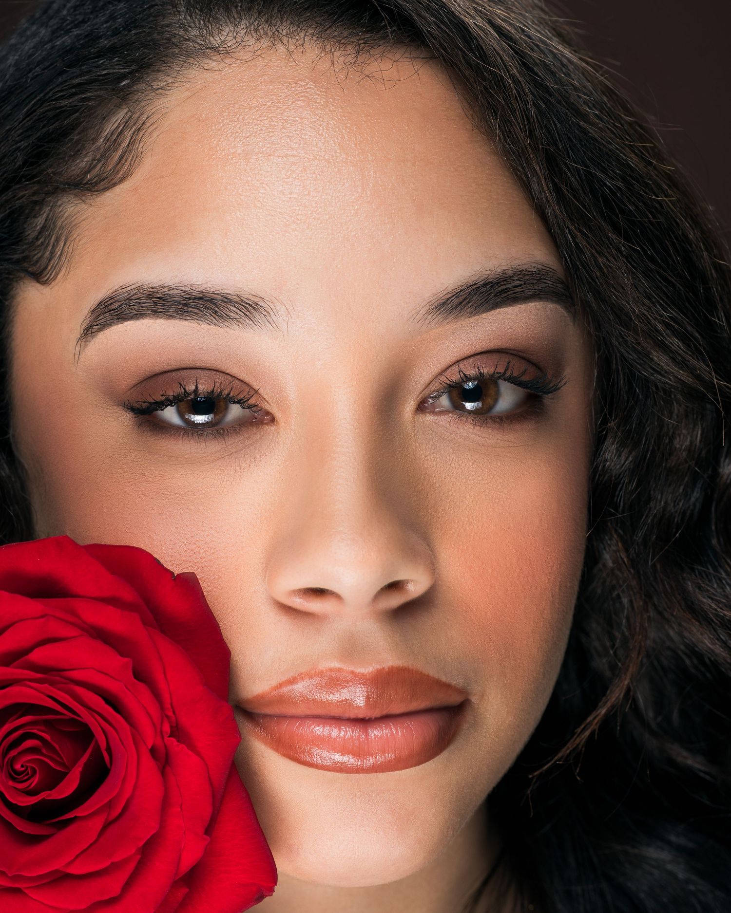 Young woman with dark hair holding a red rose, wearing natural glam makeup with defined brows and glossy lips.