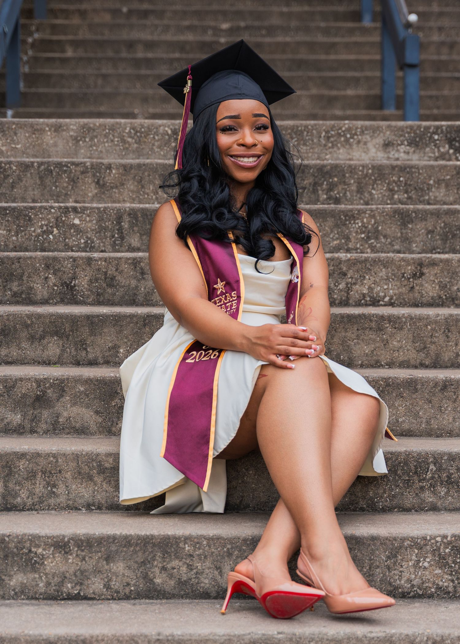 A smiling graduate in cap and gown with maroon sash sits on stone steps wearing red heels.
