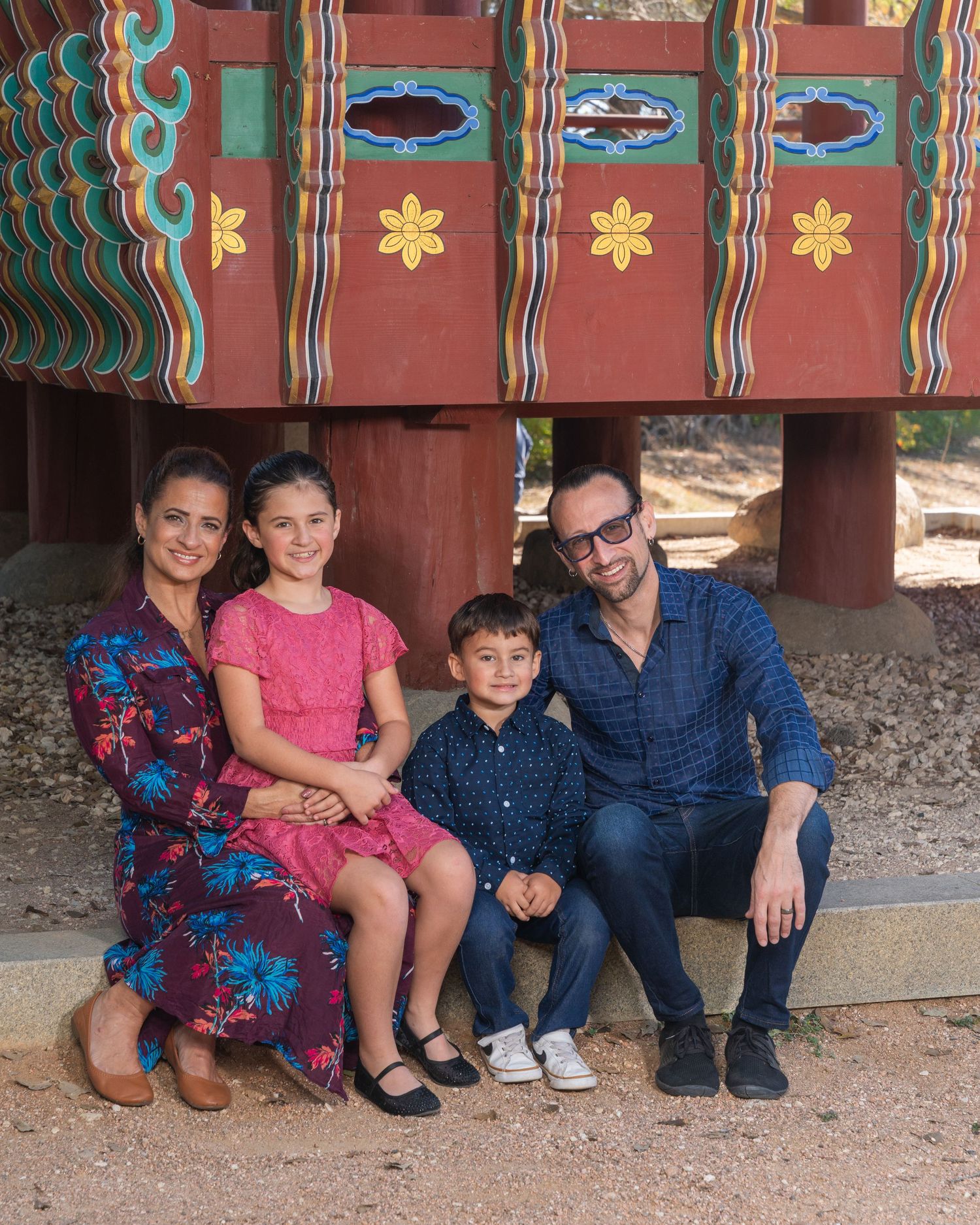 Smiling family of four poses together under vibrant traditional Korean pavilion with floral designs.