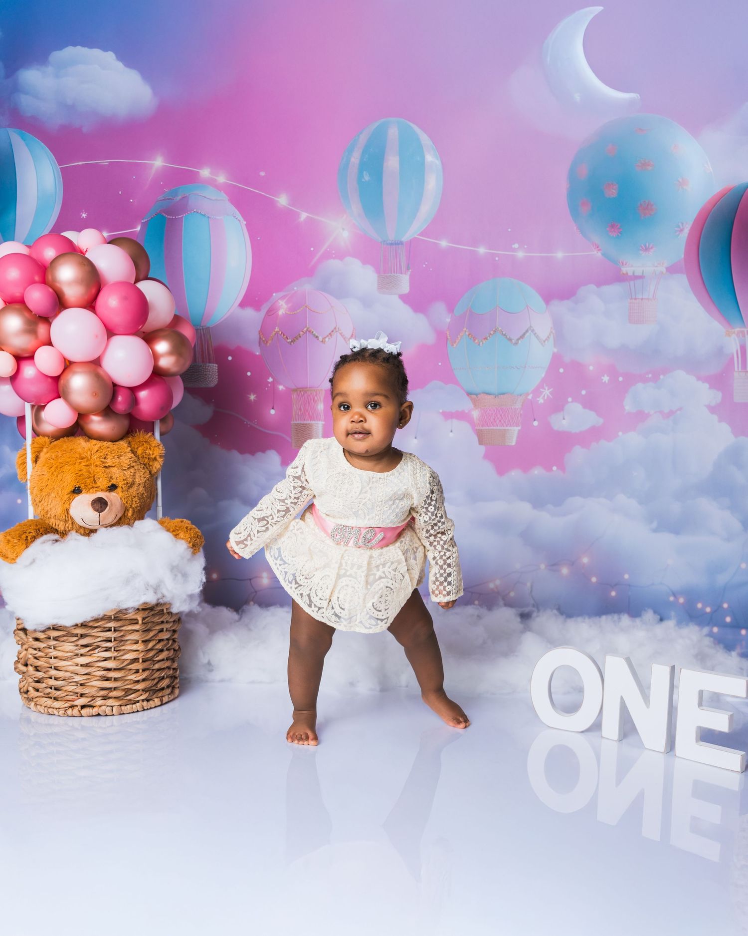 Baby girl in white lace dress stands before colorful hot air balloon backdrop celebrating her first birthday.