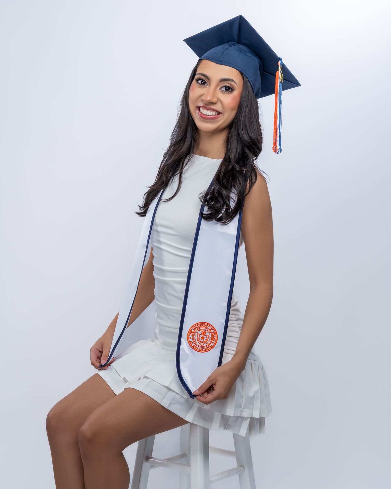 Smiling graduate in navy cap and white stole seated on stool in professional portrait.
