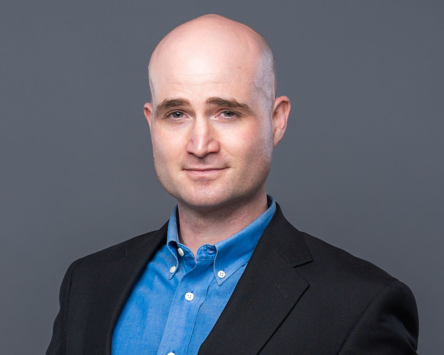 Bald professional man in blue shirt and black blazer smiling against gray background.