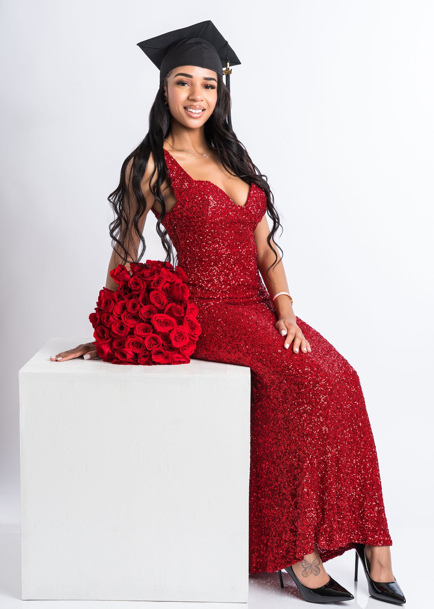 Smiling graduate in red sequin gown holding red roses bouquet wearing graduation cap on white background.