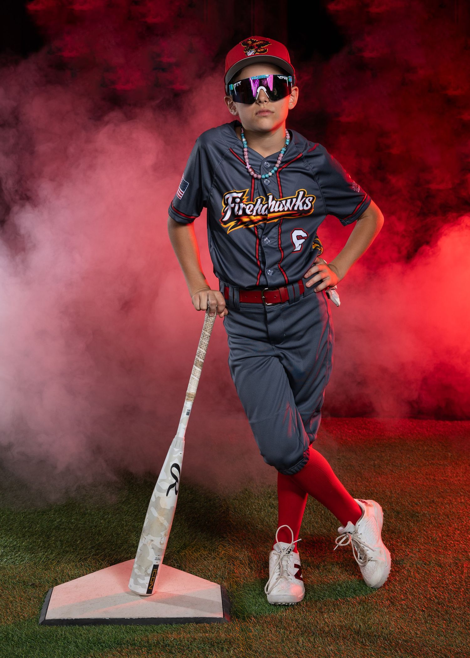 Young Firehawks baseball player in gray uniform with red socks posing with bat in dramatic red smoke.