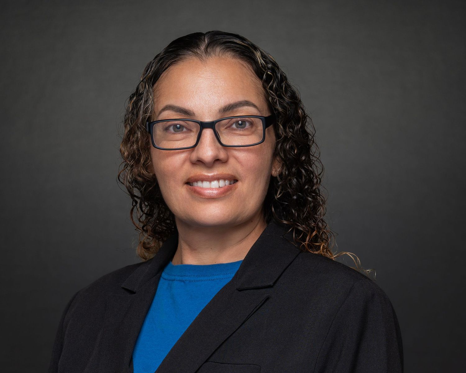 Professional headshot of a woman with curly hair and glasses wearing a blue shirt and black blazer.