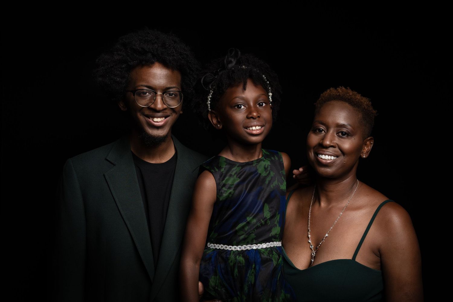 A joyful family of three poses together, featuring two adults and a child dressed in elegant attire against a dark background.