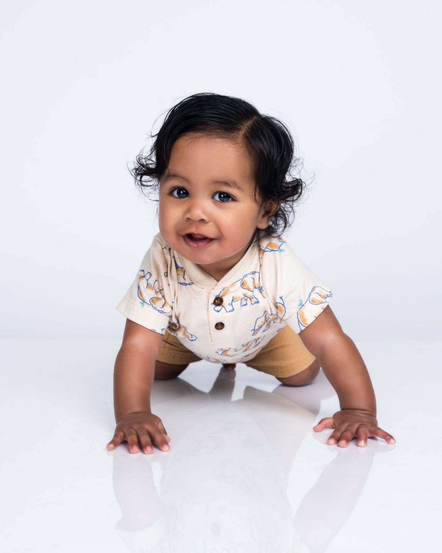 Smiling baby in a patterned cream shirt crawling on a white studio floor with dark curly hair.
