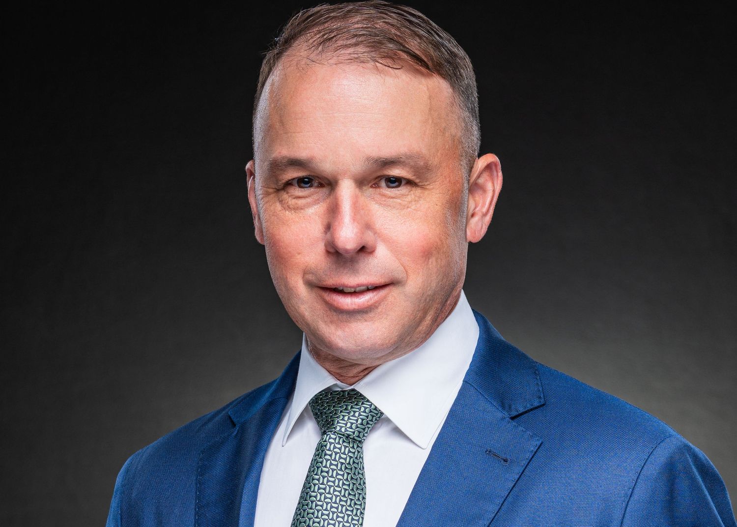 Professional headshot of a confident businessman in a blue suit and green tie against a dark background.