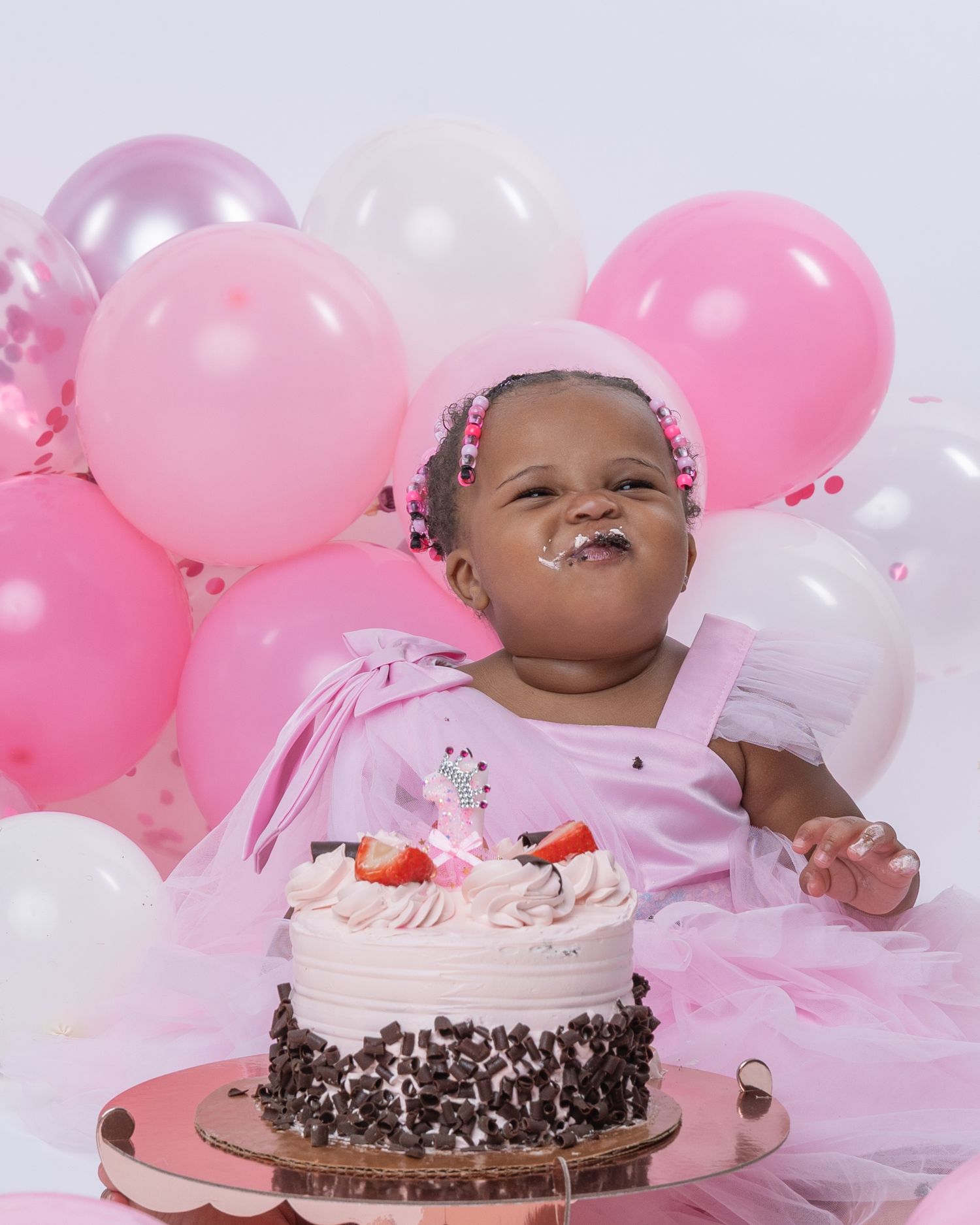 Baby girl in pink dress smashes first birthday cake surrounded by pink balloons, frosting on her smiling face.