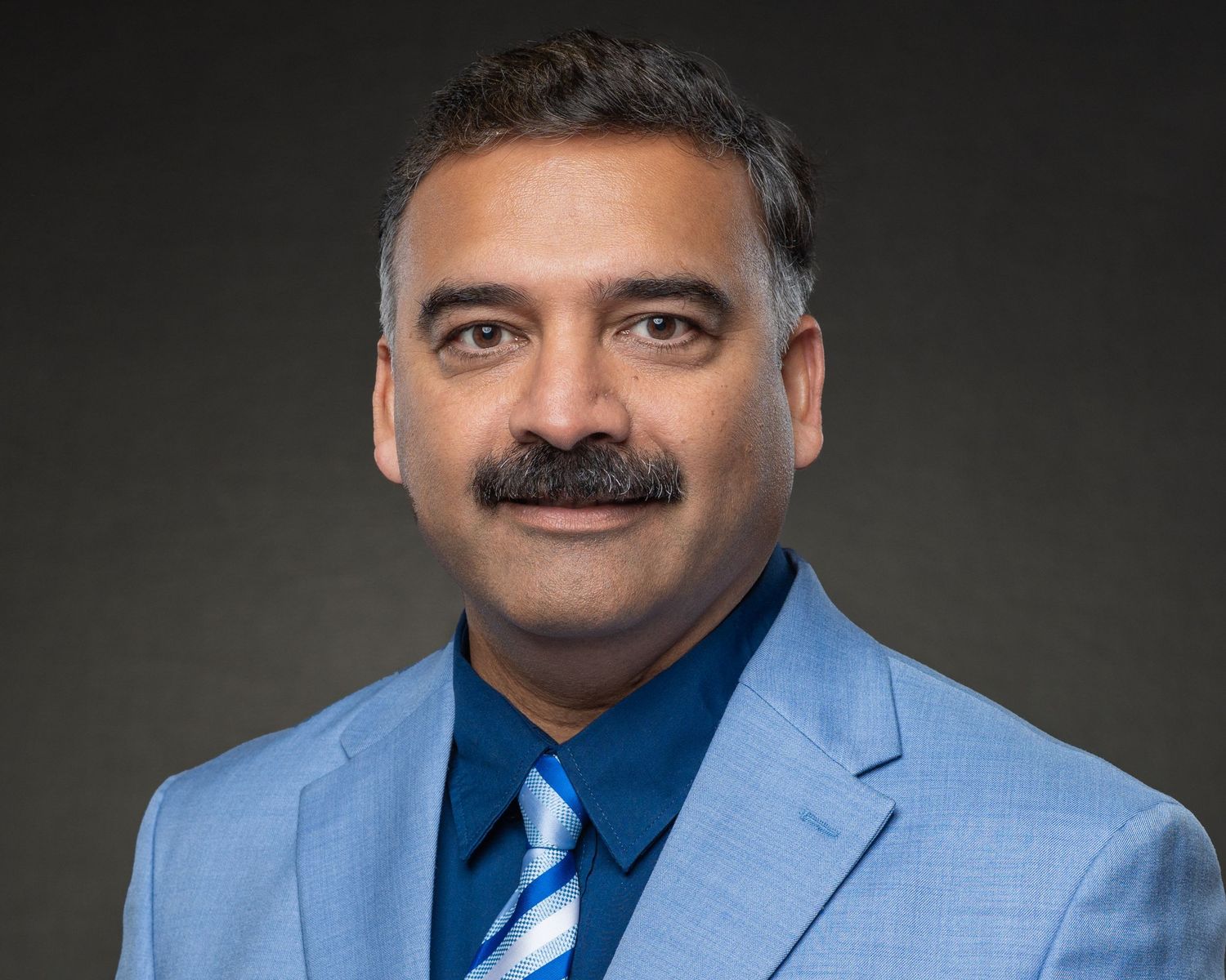 Professional headshot of a man in a light blue suit and striped tie against a gray background.