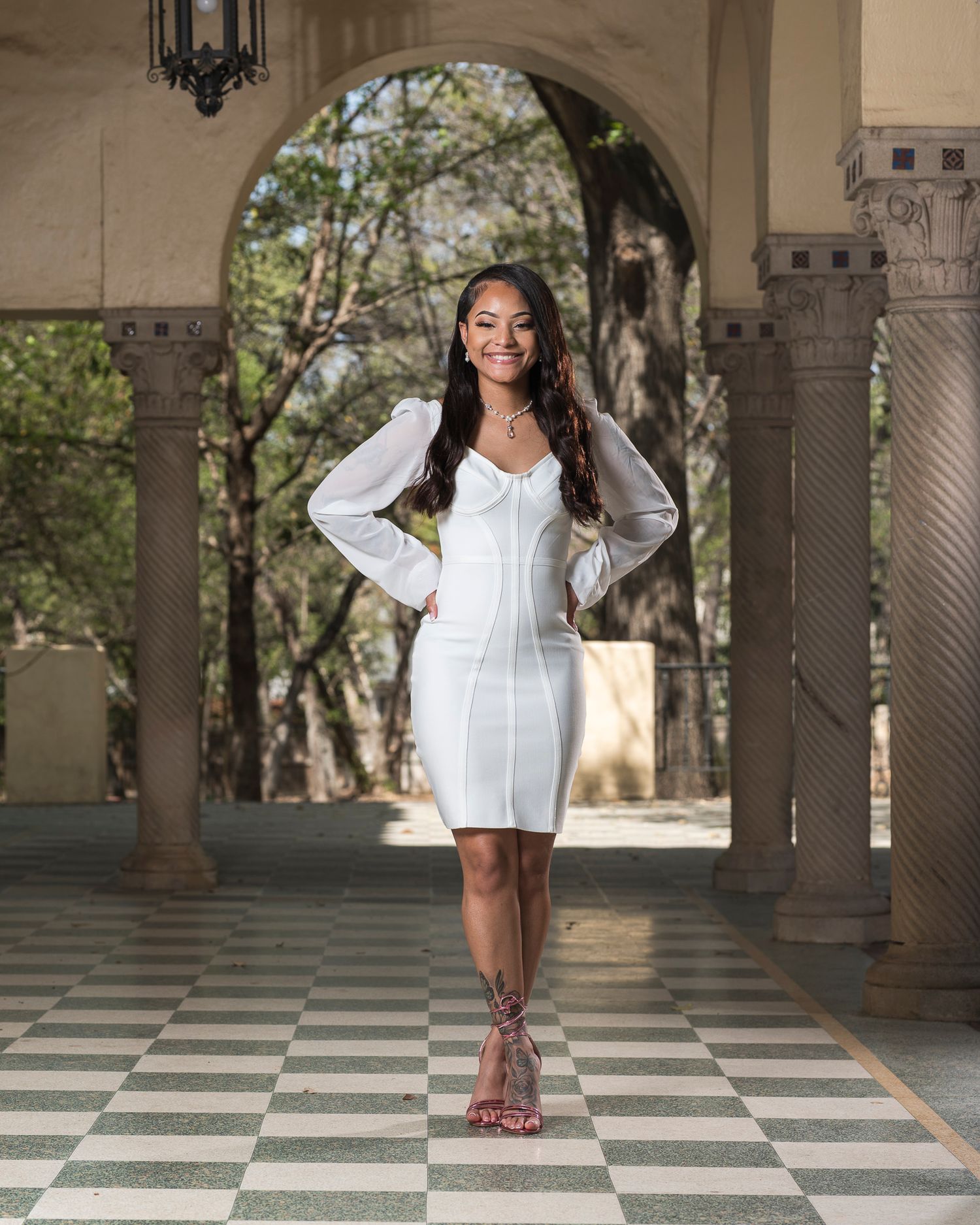 Young woman in a white long-sleeve mini dress posing under an arched colonnade outdoors.