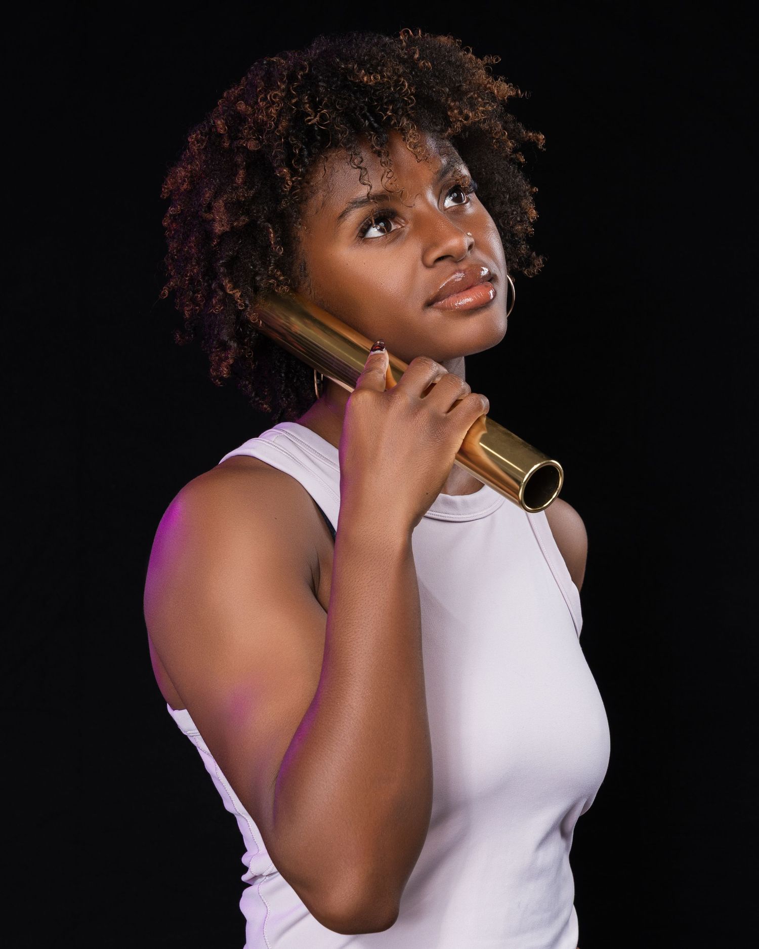 A young Black woman with natural hair holds a gold baton to her chin, gazing upward against dark background.